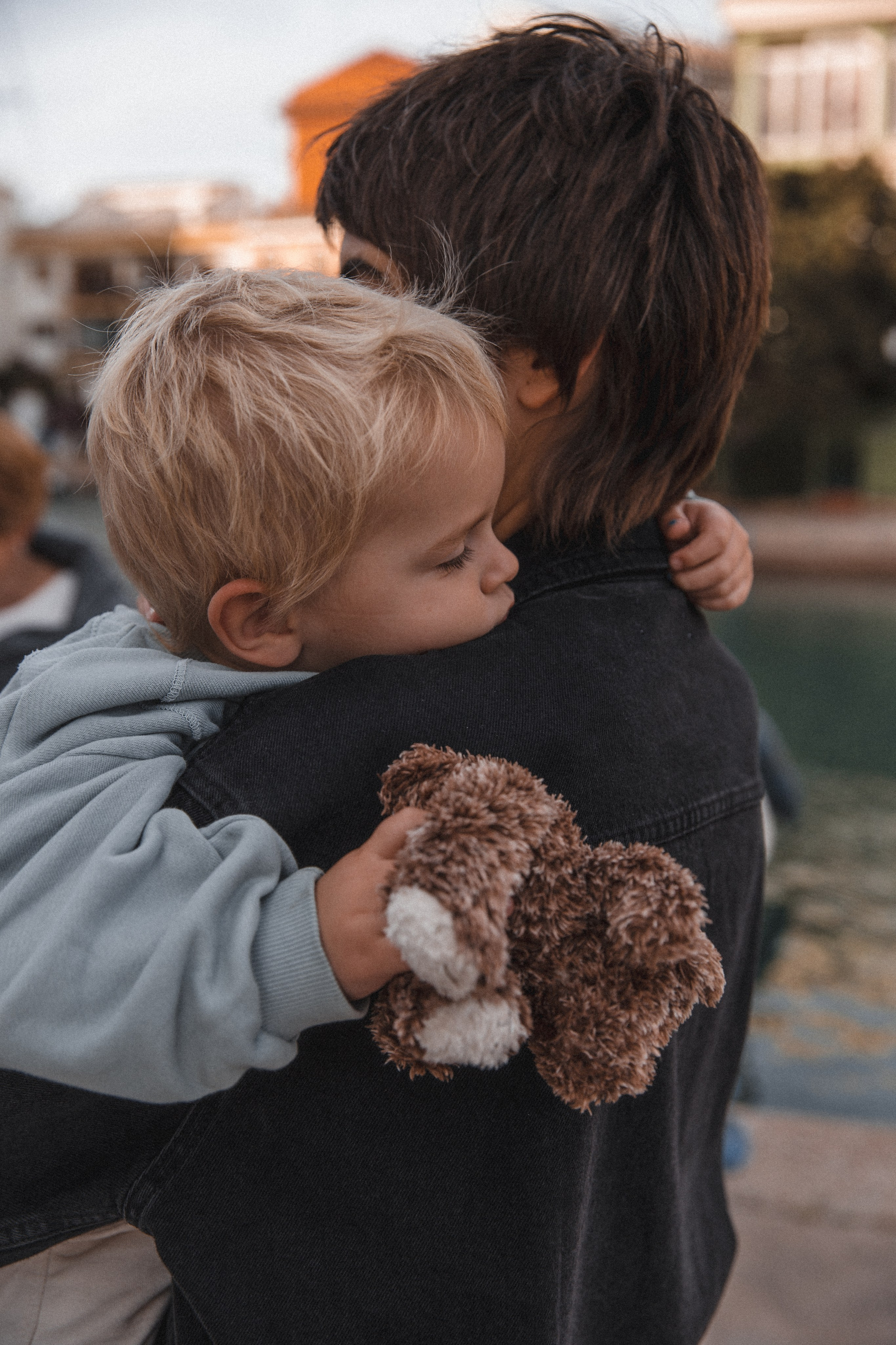 Tierno momento familiar en Valencia, España — niño abrazando su peluche mientras descansa sobre el hombro de su madre junto al agua.