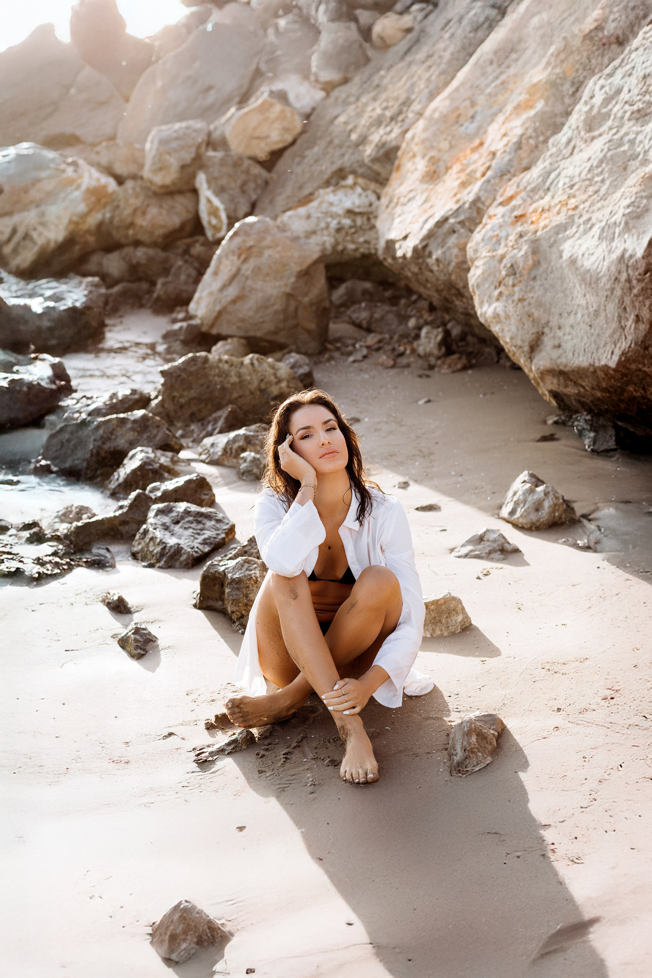 Атмосферная портретная фотосессия на пляже в Таррагоне, Испания — девушка босиком в белой рубашке сидит среди скал у побережья, в лучах мягкого естественного света. Прекрасный выбор для любителей художественной и чувственной съёмки в Таррагоне и живописных уголках Испании.