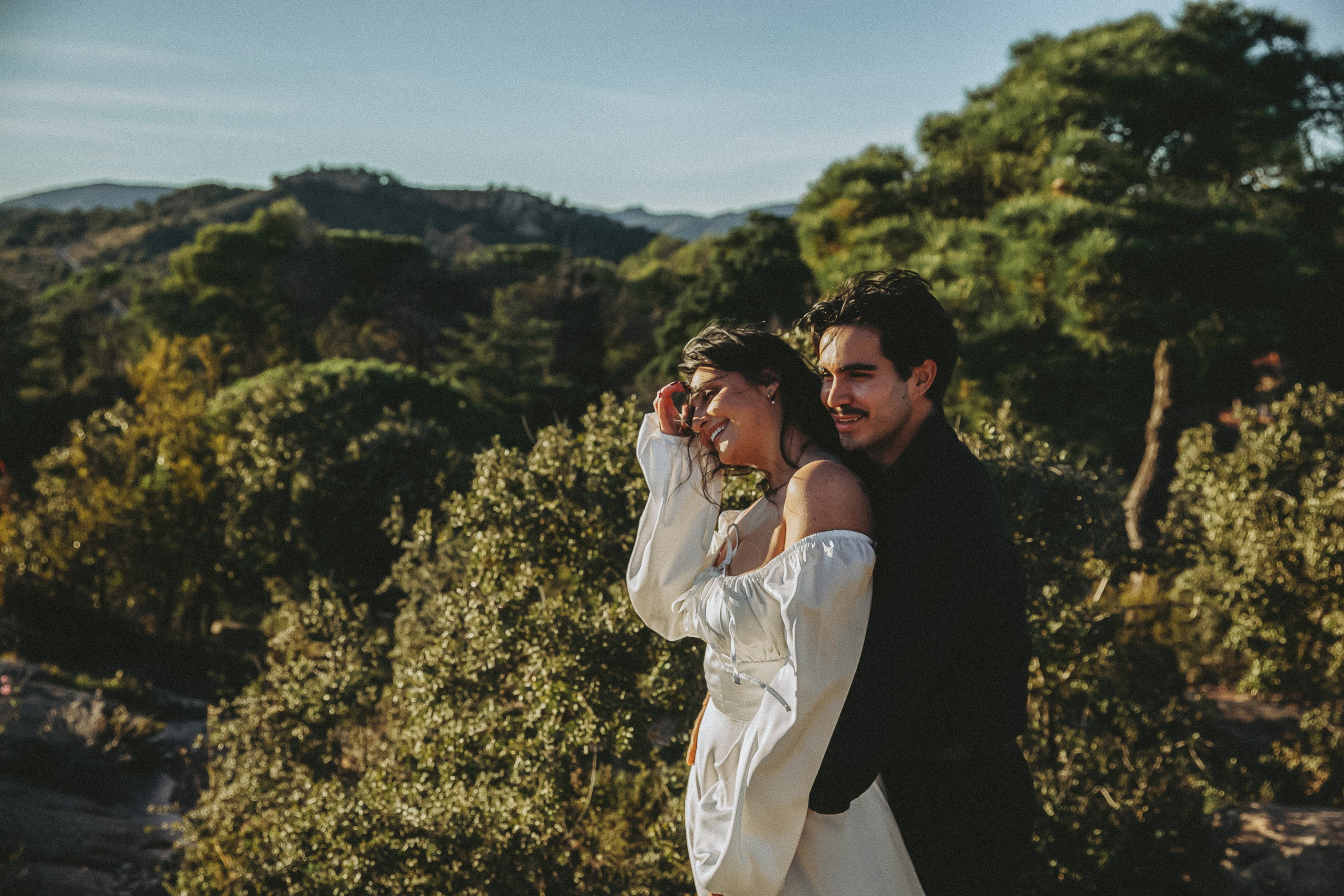 Engagement photoshoot in València, Spain, featuring a romantic couple embracing in lush Mediterranean nature with rolling green hills in the background, captured in warm golden-hour light — a natural and emotional engagement and wedding love story image ideal for engagement photography, pre-wedding sessions, elopements, and professional wedding photoshoots in València and across Spain.