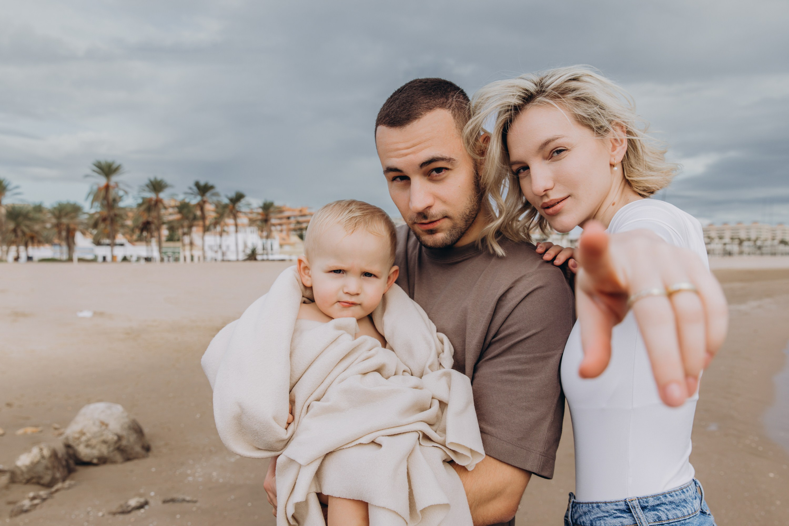 Sesión de fotos familiar divertida y moderna en la playa de Valencia, España — padres con estilo posan con confianza junto a su bebé envuelto en una manta, capturando un momento audaz y lleno de amor junto al mar. Ideal para quienes buscan sesiones familiares creativas y naturales en Valencia y en toda España.