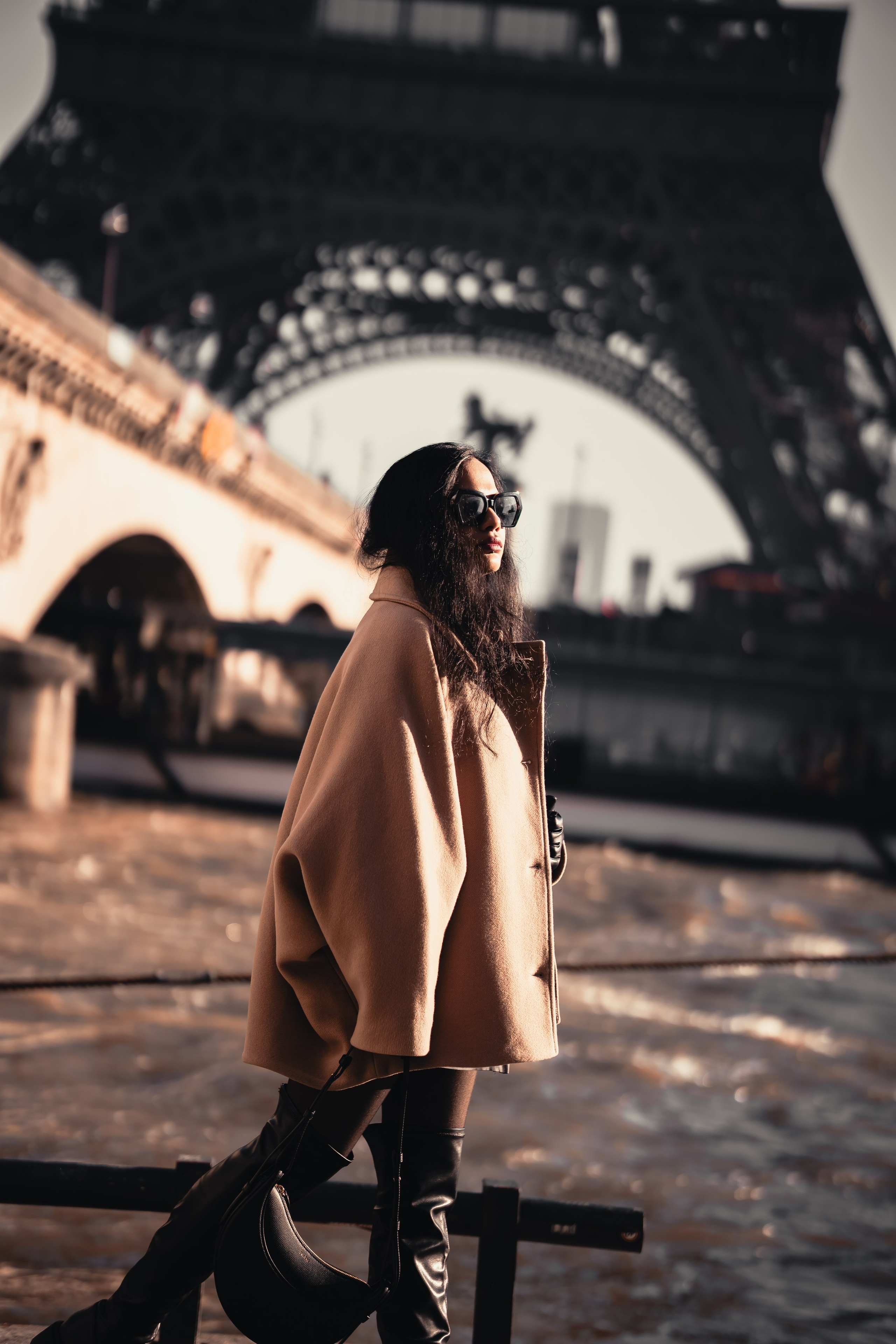 a woman wearing oversized brown jacket and standing with paris bridge and eiffel tower view behind her
