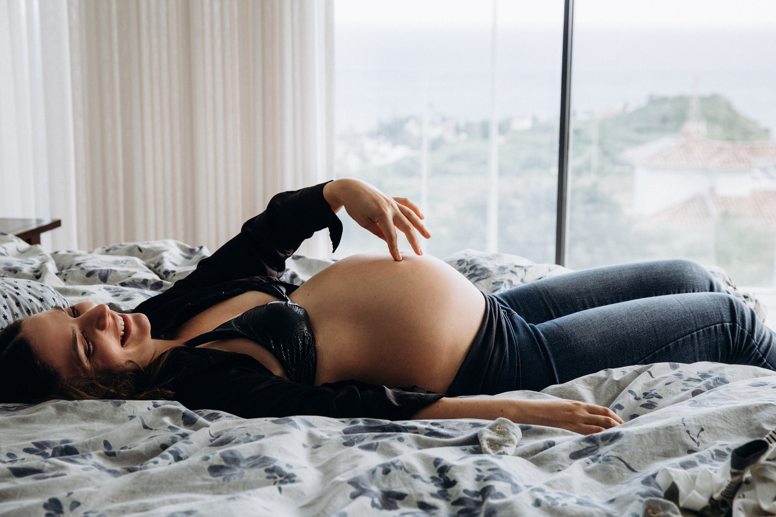 Sesión de fotos en interior en Madrid, España — mujer embarazada riendo acostada en una cama con lencería, mostrando belleza natural y conexión íntima durante el embarazo. Ideal para quienes buscan sesiones relajadas y elegantes en Madrid.