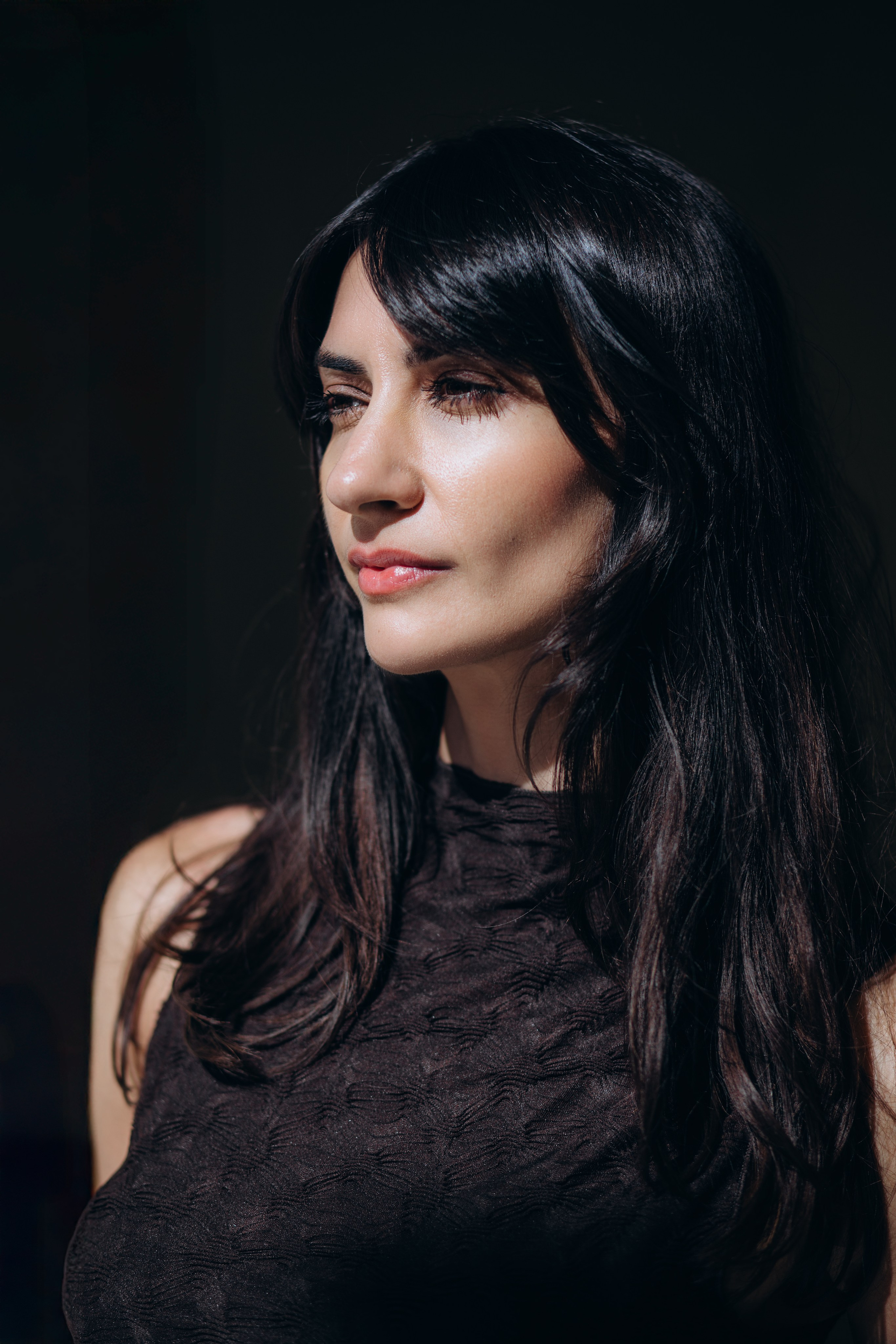Крупный портрет женщины с длинными темными волосами и естественным макияжем в драматичном свете на темном фоне — минималистичная и выразительная портретная фотография в Барселоне, Испания, идеально подходит тем, кто ищет элегантные и художественные портретные фотосессии в Барселоне и Испании.