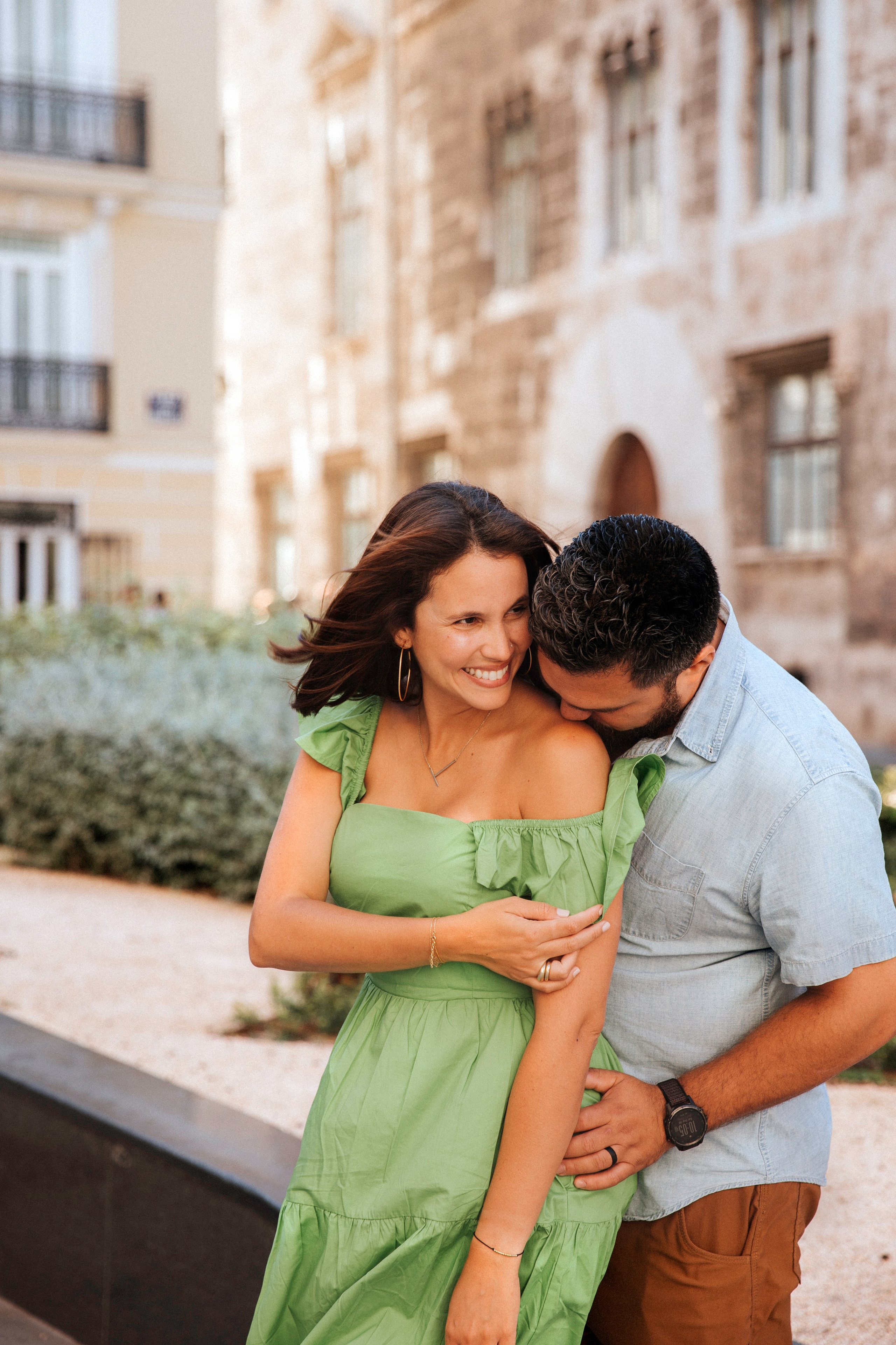 Playful couple sharing a joyful moment in the historic center of Valencia, Spain, as the man lovingly leans into his partner’s shoulder while she smiles in a vibrant green dress.