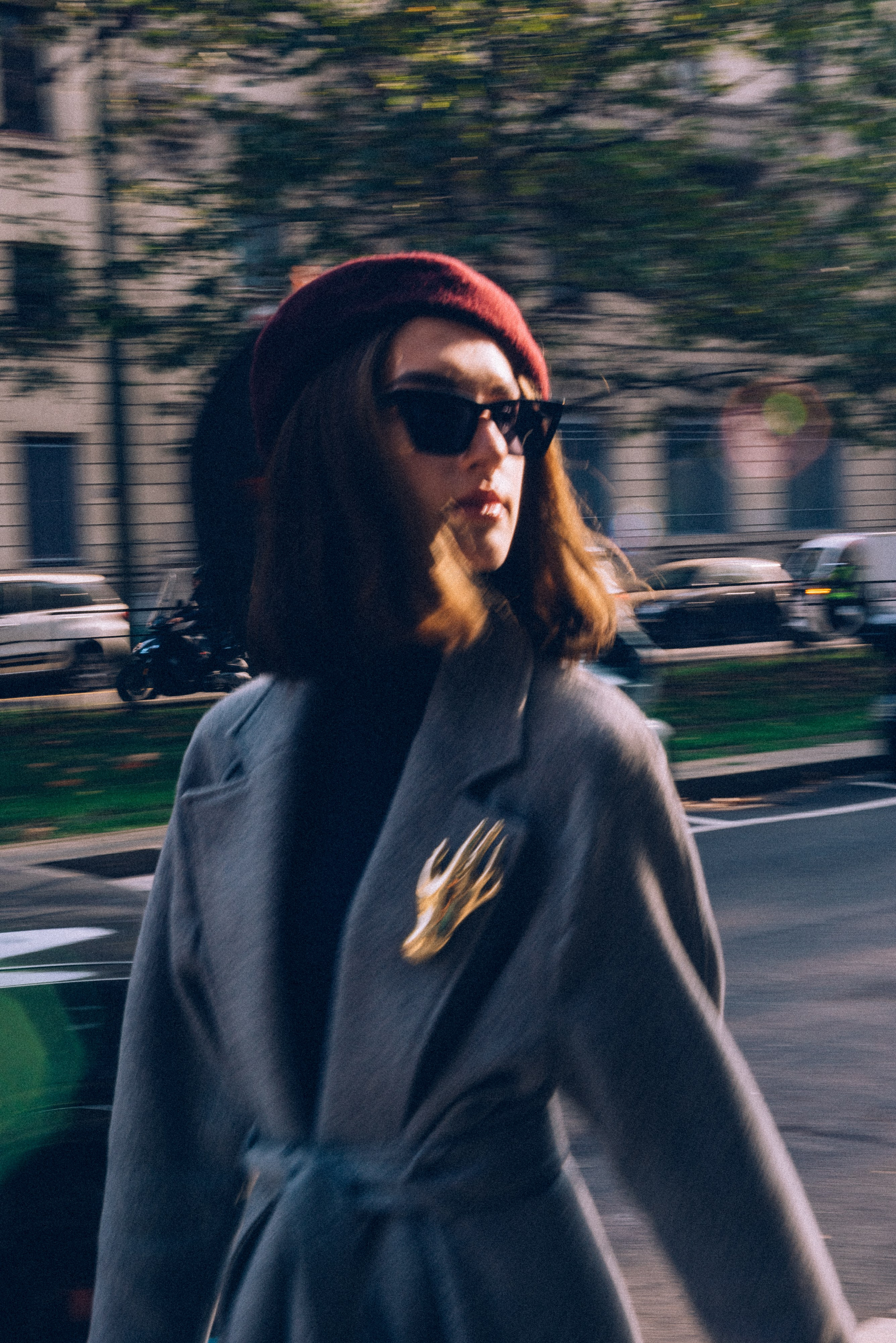Stylish woman in a gray coat with a vintage gold brooch and burgundy beret walking through Milan's vibrant streets, blending fashion and urban energy