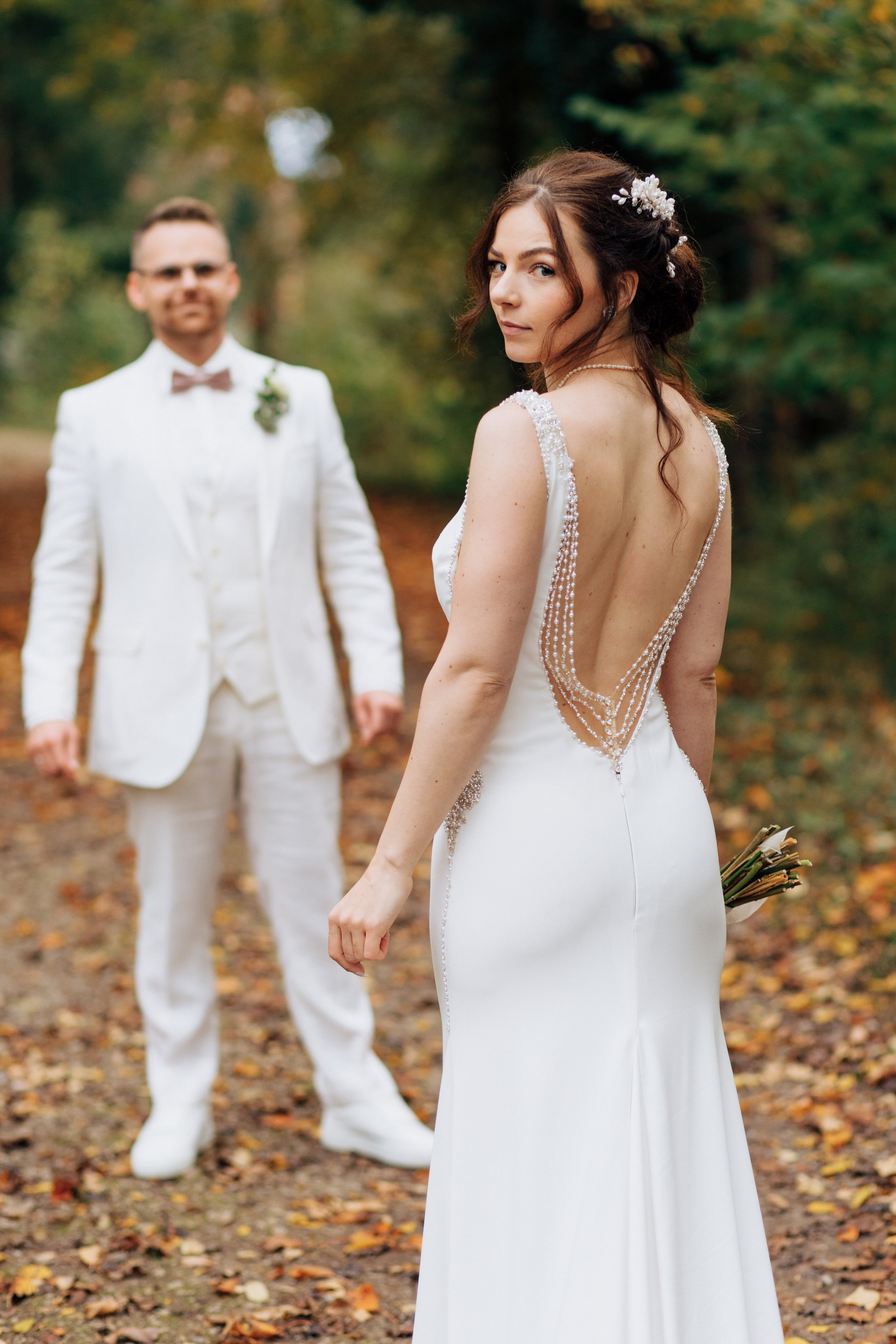 Die Braut steht in ihrem eleganten, figurbetonten Hochzeitskleid, während der Bräutigam sie anlächelt.