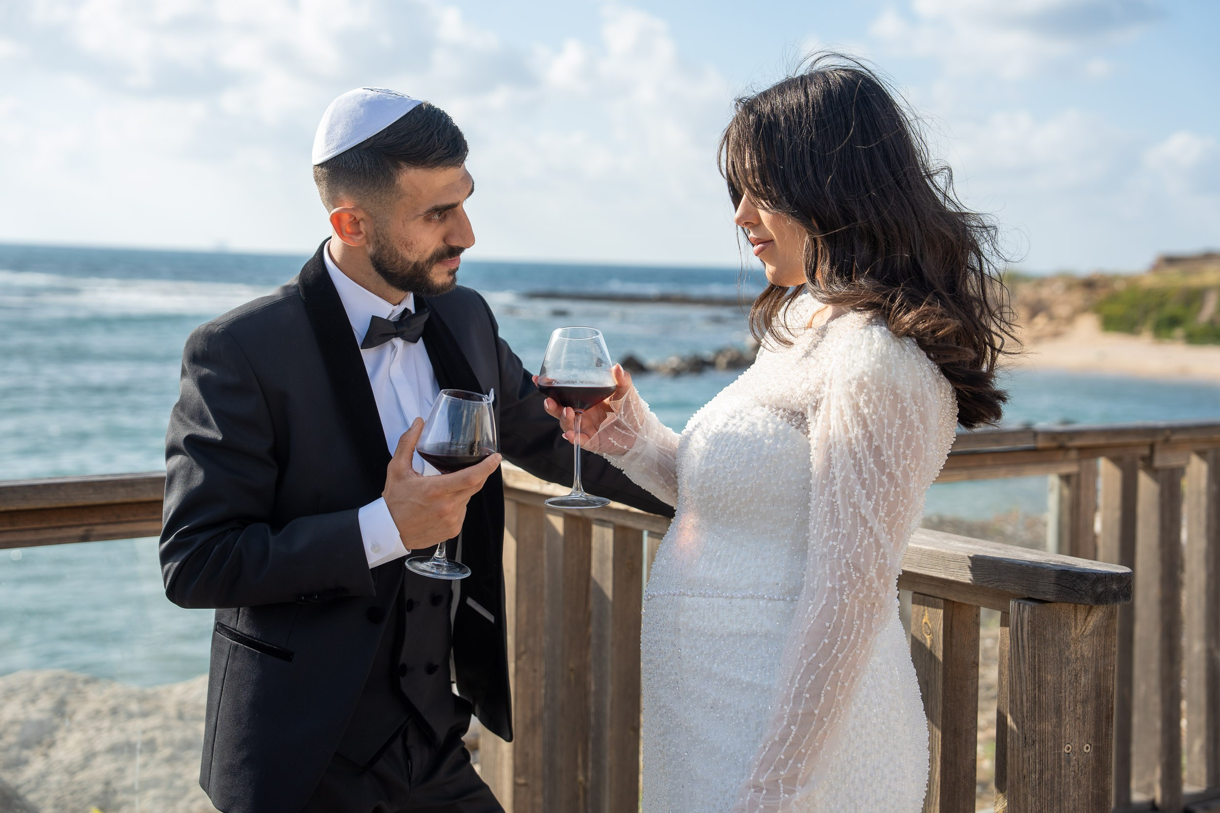 Bride Natali and groom Shimon holding wine glasses by the sea during their wedding photoshoot in Caesarea, Israel