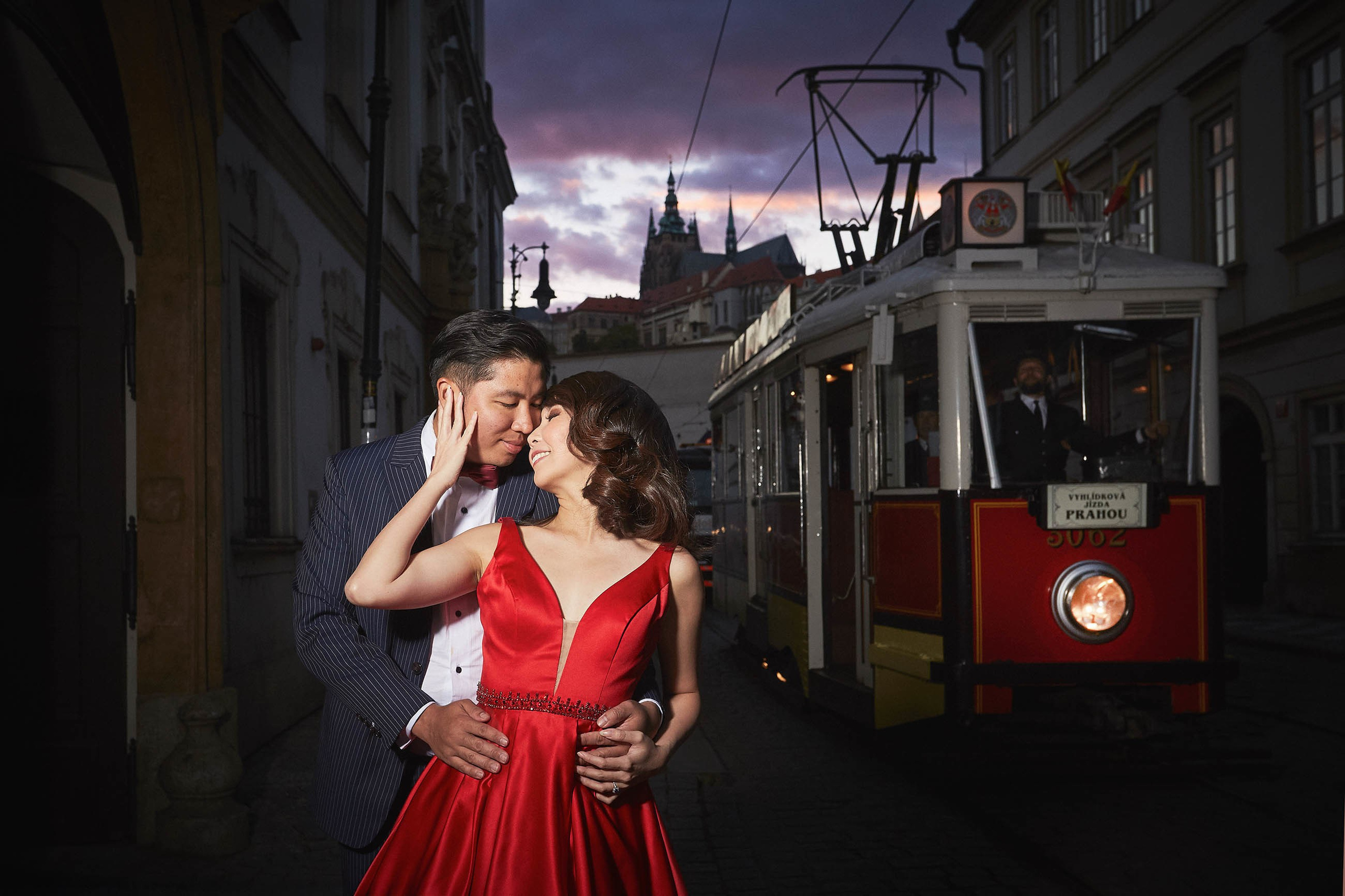 Asian woman in vibrant red dress is embraced from behind by her partner as a historical tram pulls up behind them at dusk as St. Vitus Cathedral looms above them in Prague