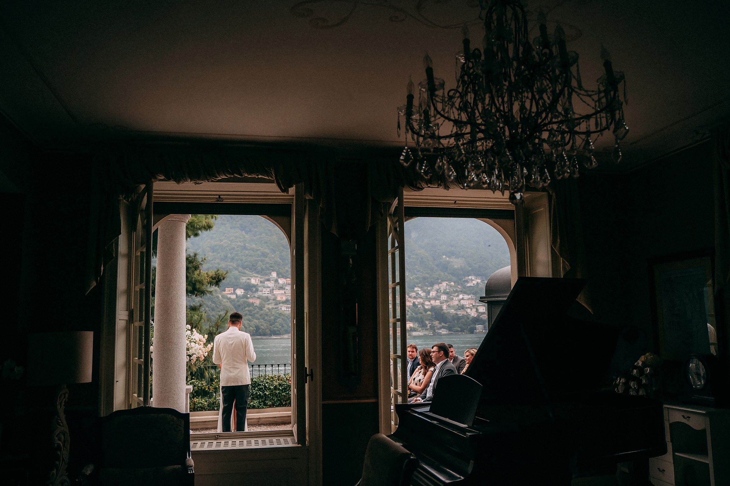 Elegant lakeside villa wedding with bride in white jacket addressing guests from balcony overlooking scenic hillside town.