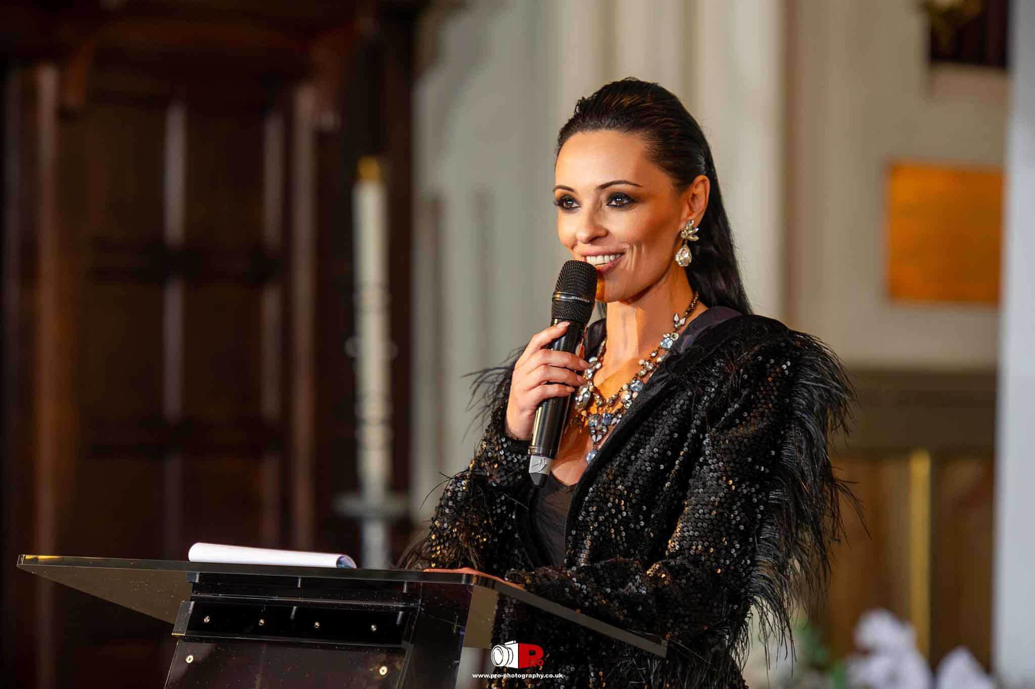 A woman in a sparkling black gown is hosting a fashion gala with a microphone in hand.