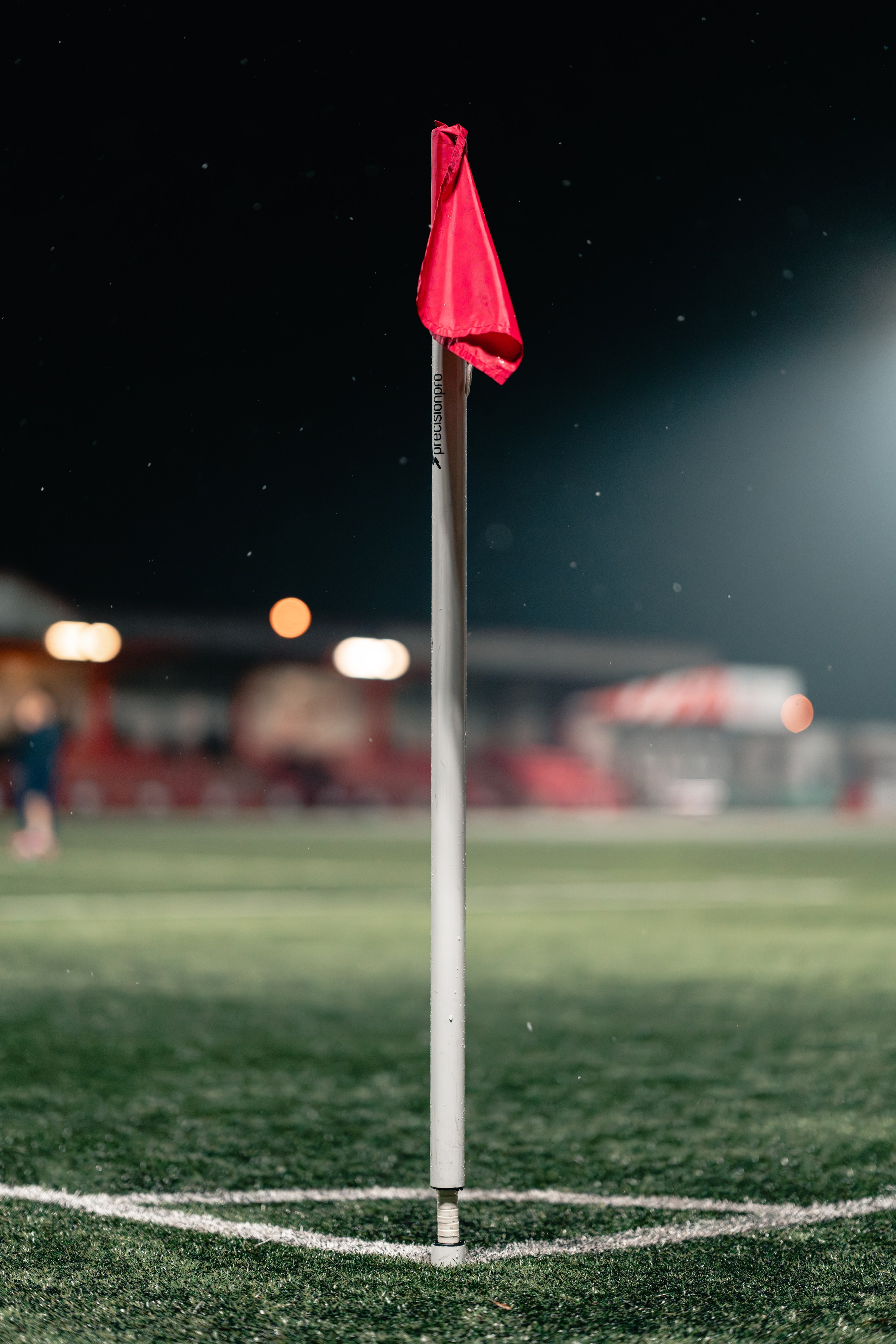 Red corner flag under floodlights with raindrops visible on a night-time pitch