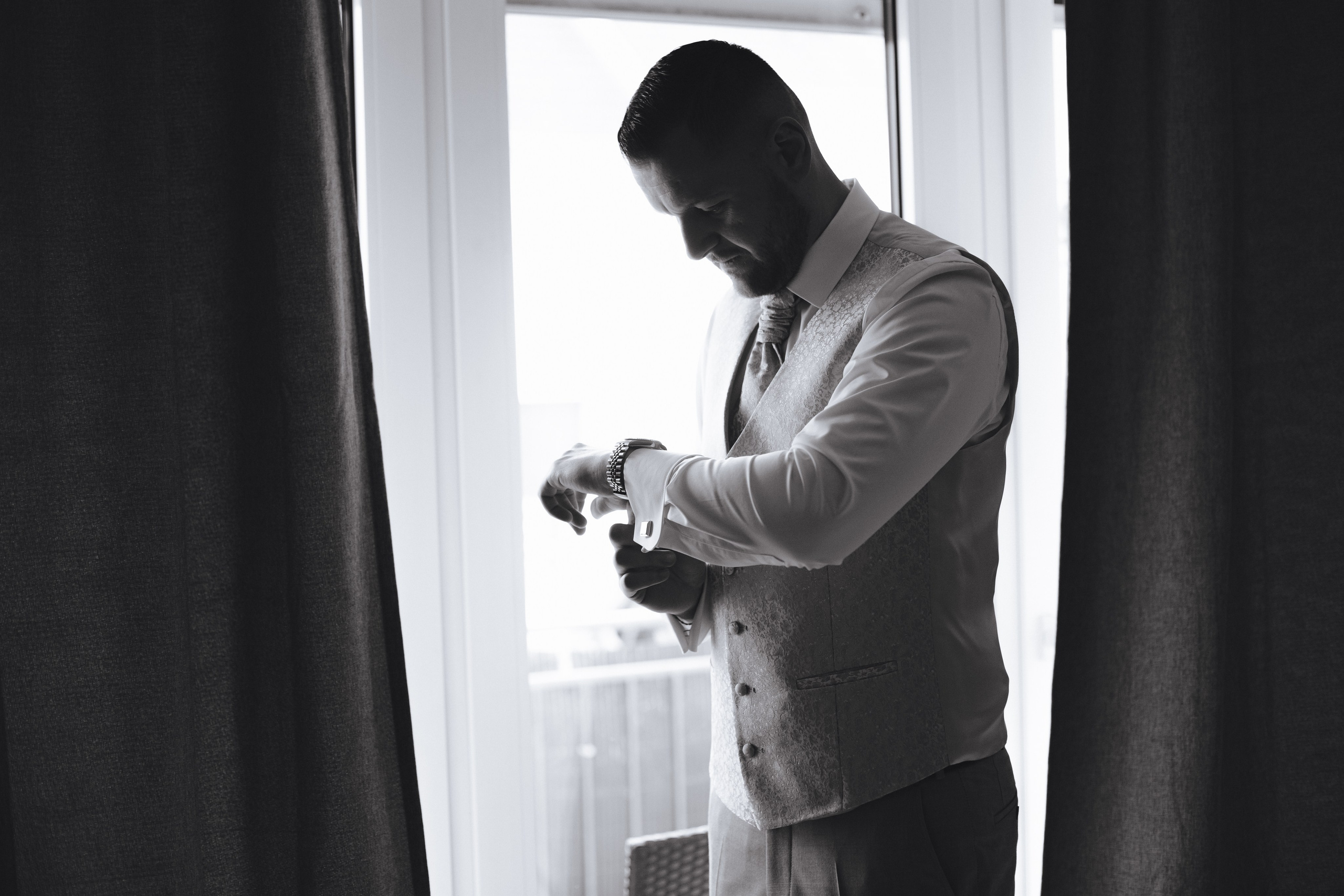 Der Bräutigam steht am Fenster und richtet seine Manschettenknöpfe und Uhr. Ein eleganter Moment der Hochzeitsvorbereitung.