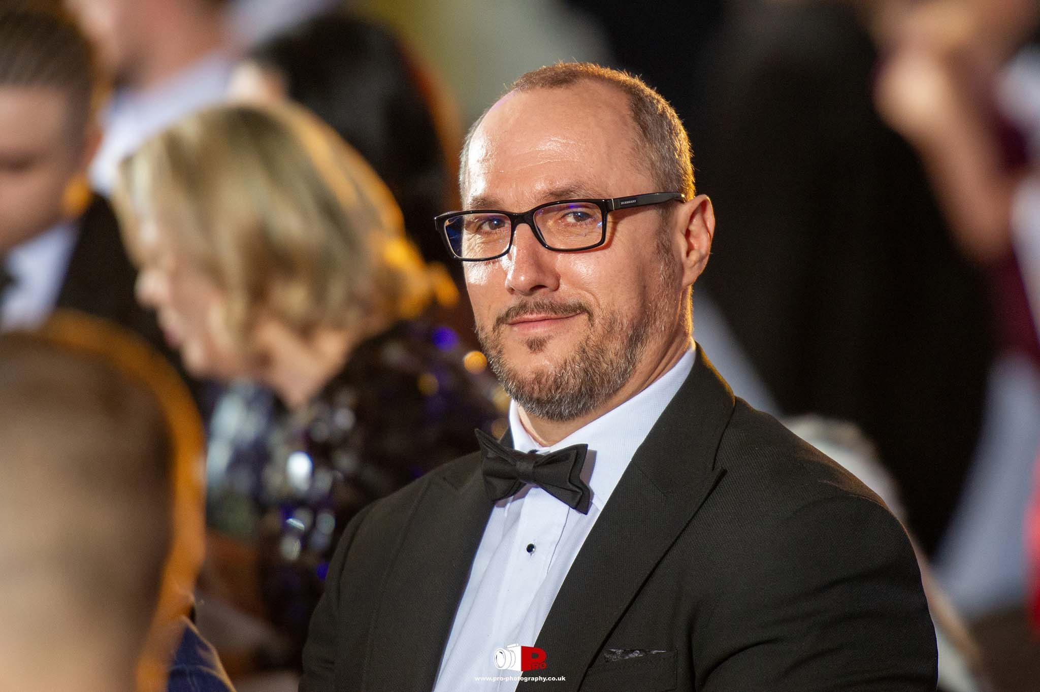 A distinguished man in glasses and a tuxedo smiling at a formal gala dinner event.