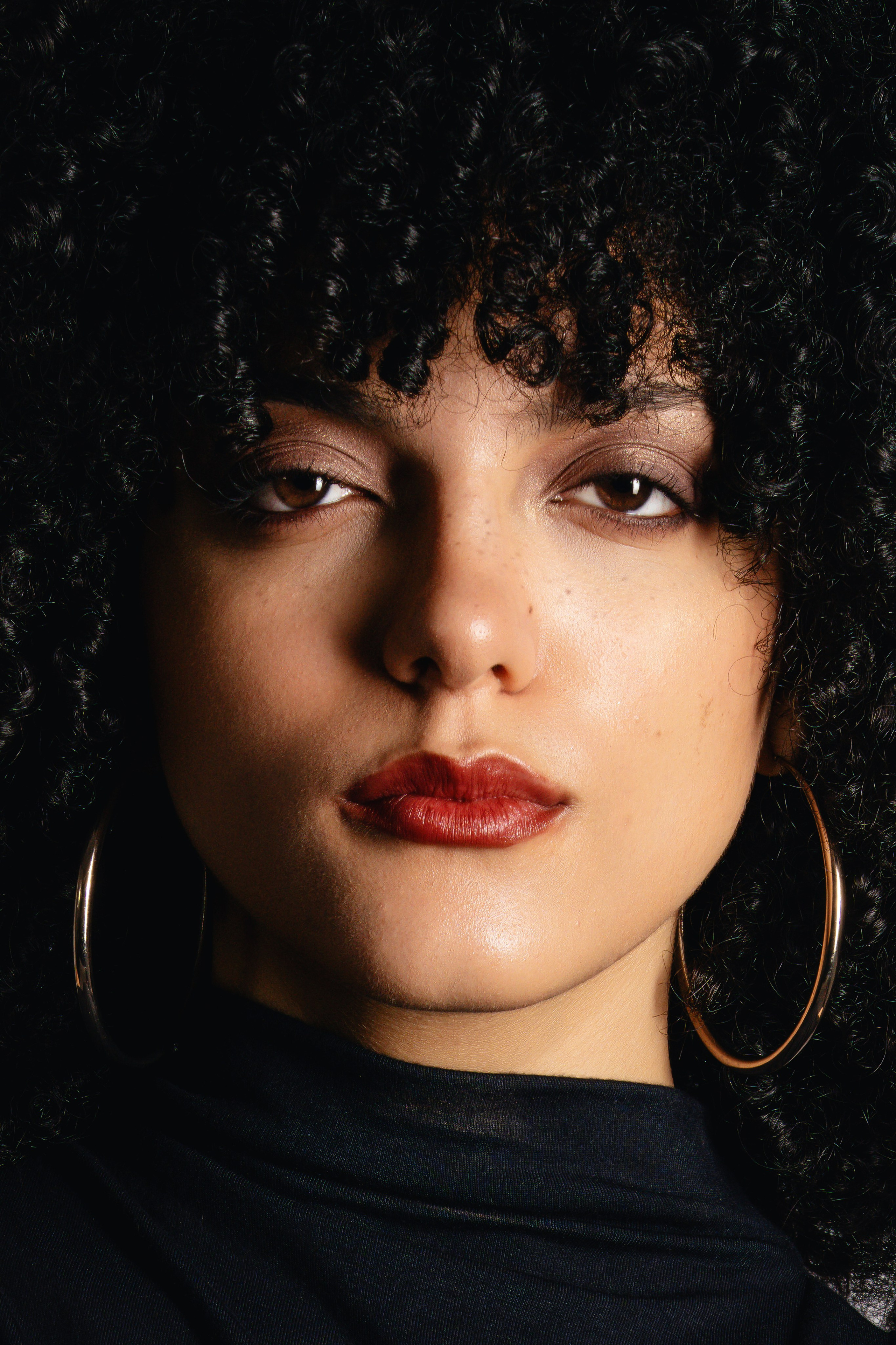 Fashion portrait of a woman with dark curly hair and hoop earrings, photographed in a clean editorial style for high-end portrait and luxury brand imagery.