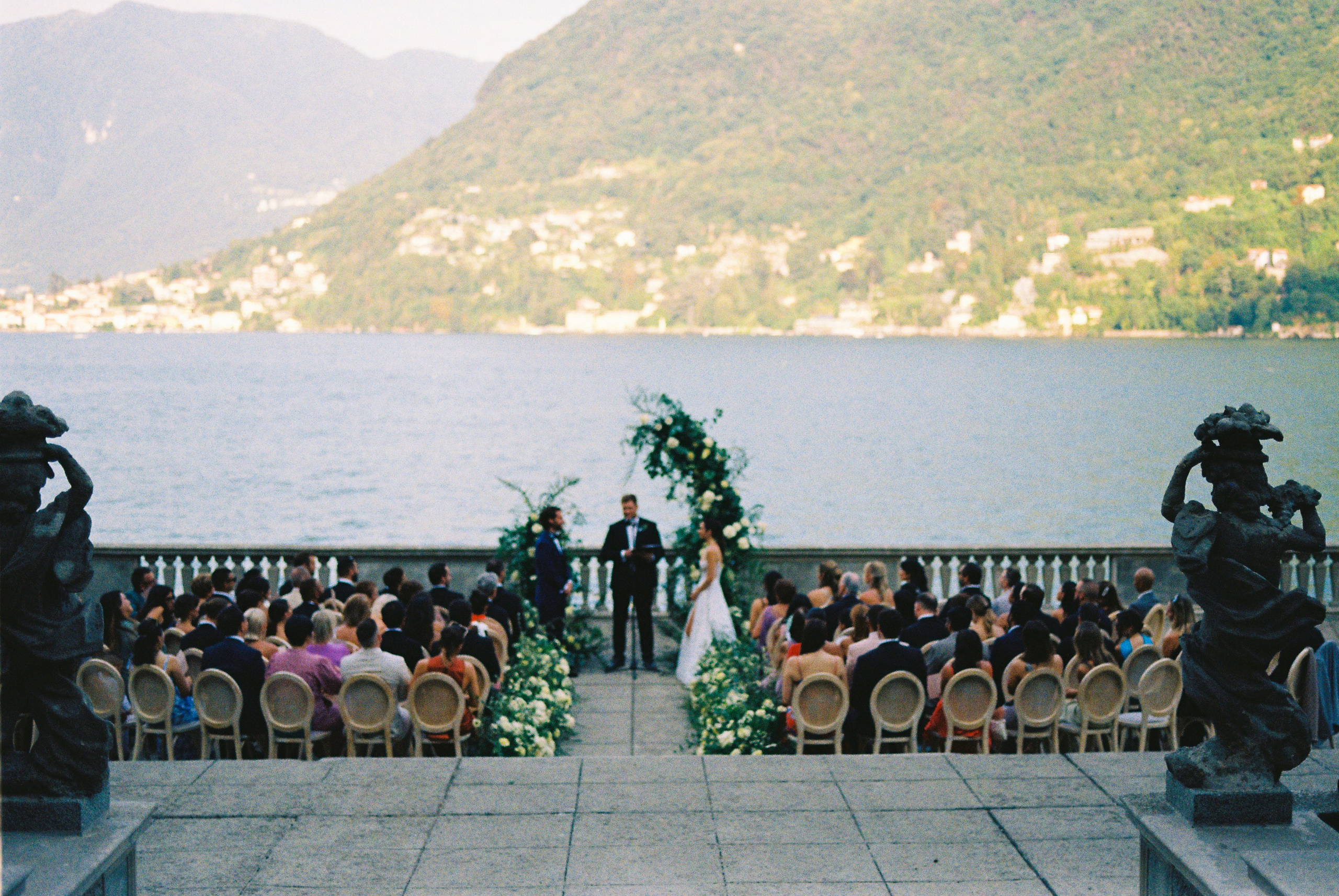 Bride and groom exchange vows by lake, with guests seated against a scenic mountain backdrop.