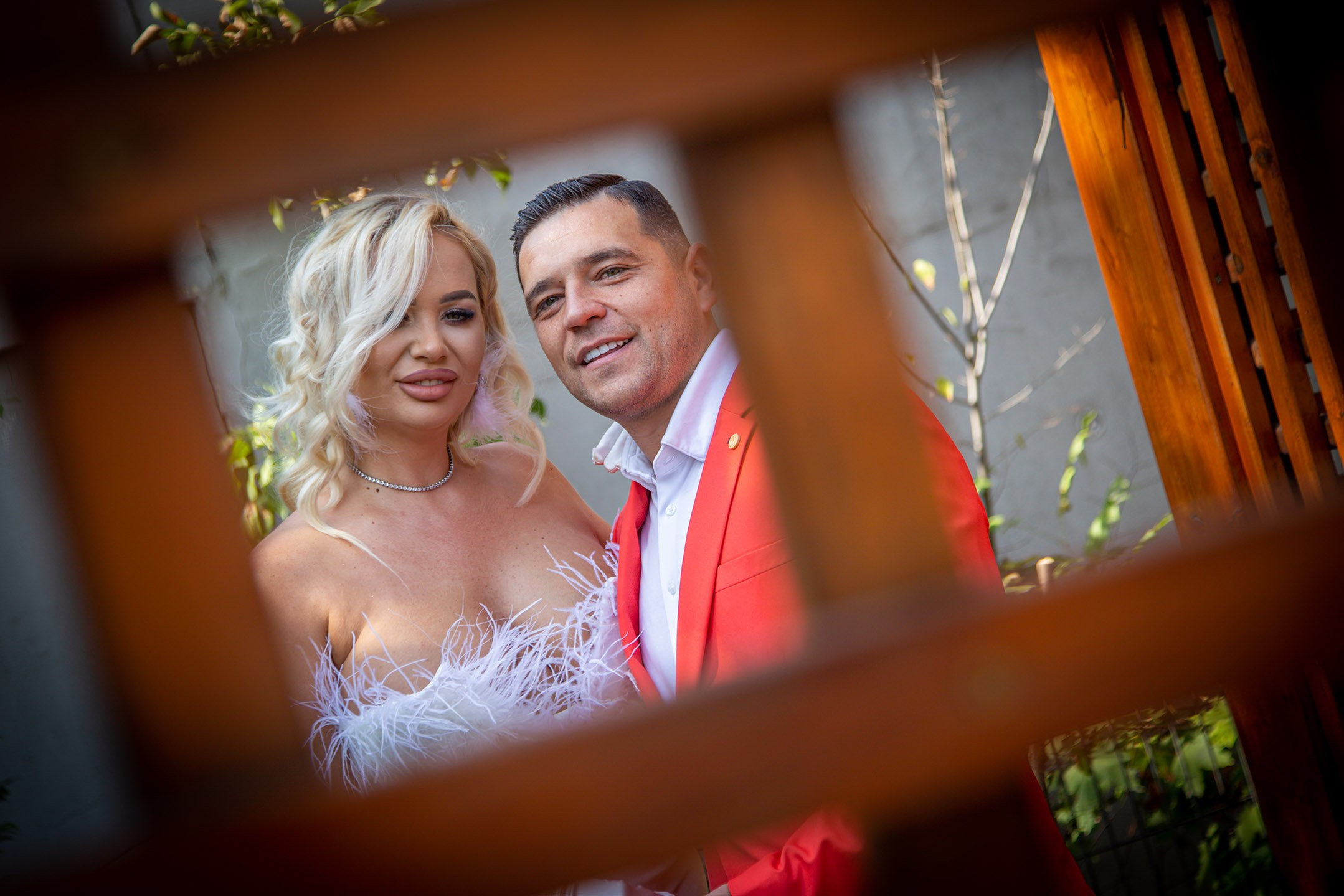 Cuplu de miri la ședința foto – mireasa în rochie albă cu pene și mirele în sacou roșu, poză romantică în aer liber