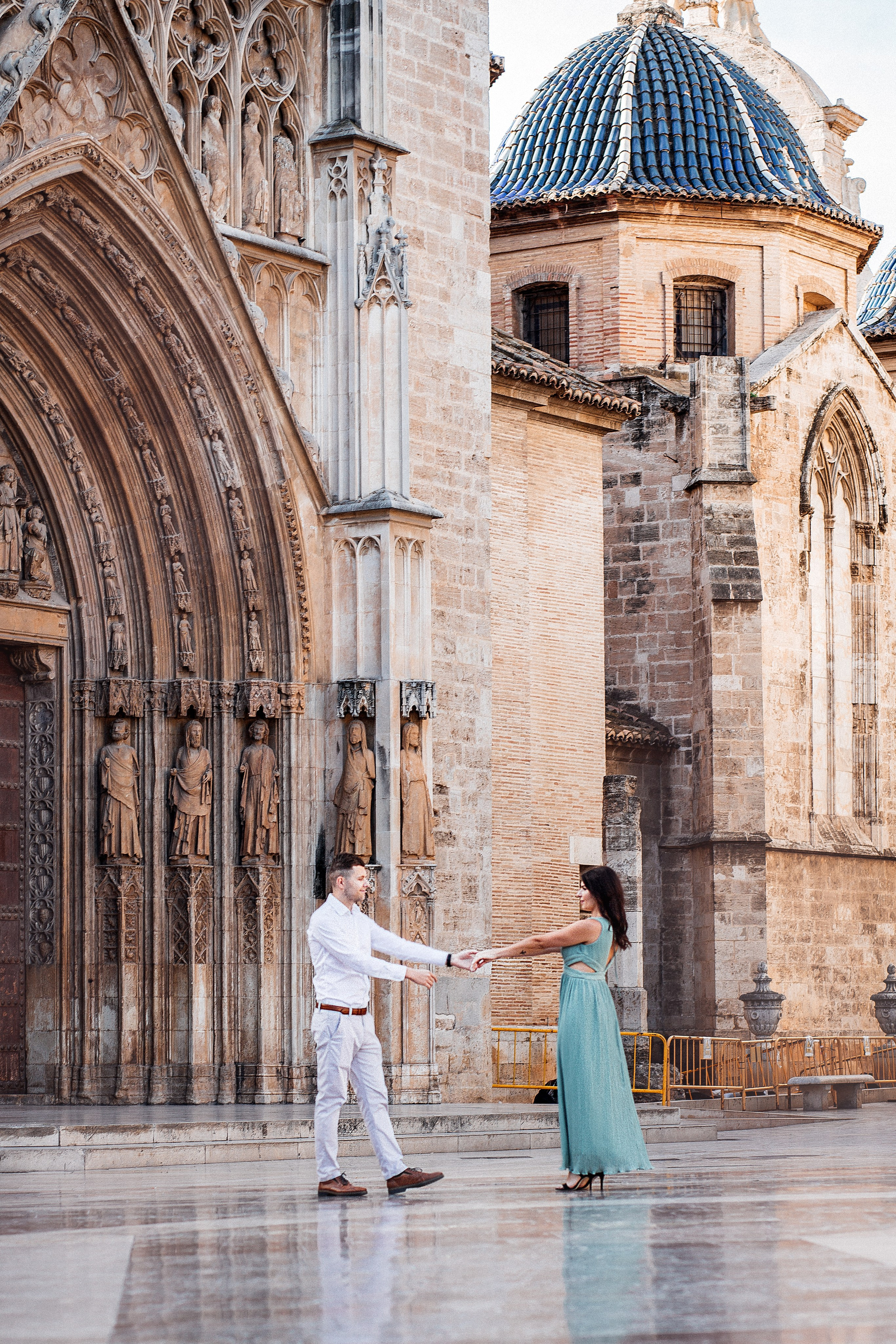Romantic love story photo of a couple holding hands and smiling at each other in front of the grand entrance of Valencia Cathedral, surrounded by historic gothic architecture and soft golden light.