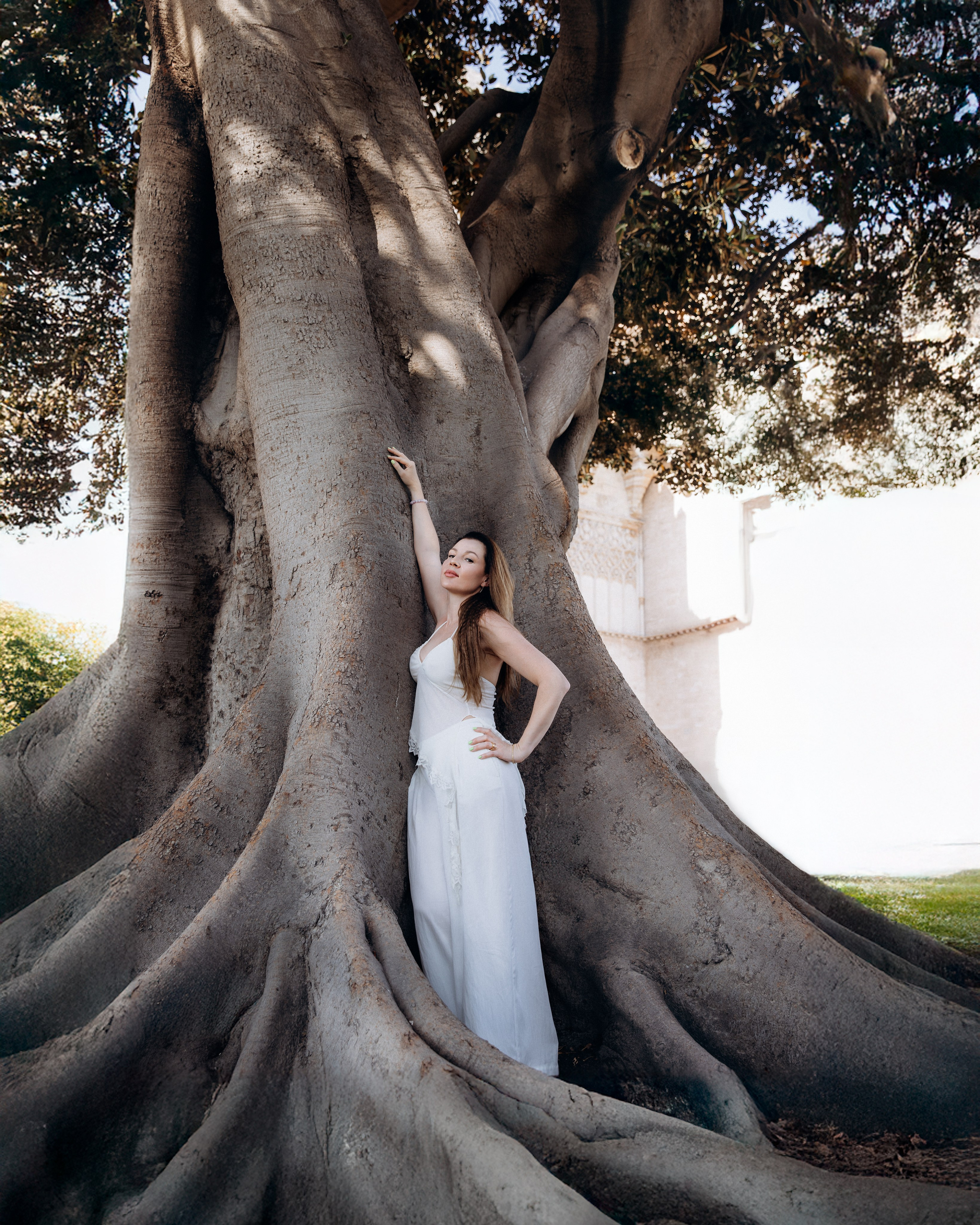 Женская фотосессия в Валенсии, Испания, с элегантным уличным портретом женщины в струящемся белом платье рядом с величественным древним деревом с мощными корнями, снятым в мягком естественном свете — идеально для женской портретной фотографии, художественных уличных съёмок и профессиональных женских фотосессий в Валенсии и по всей Испании.