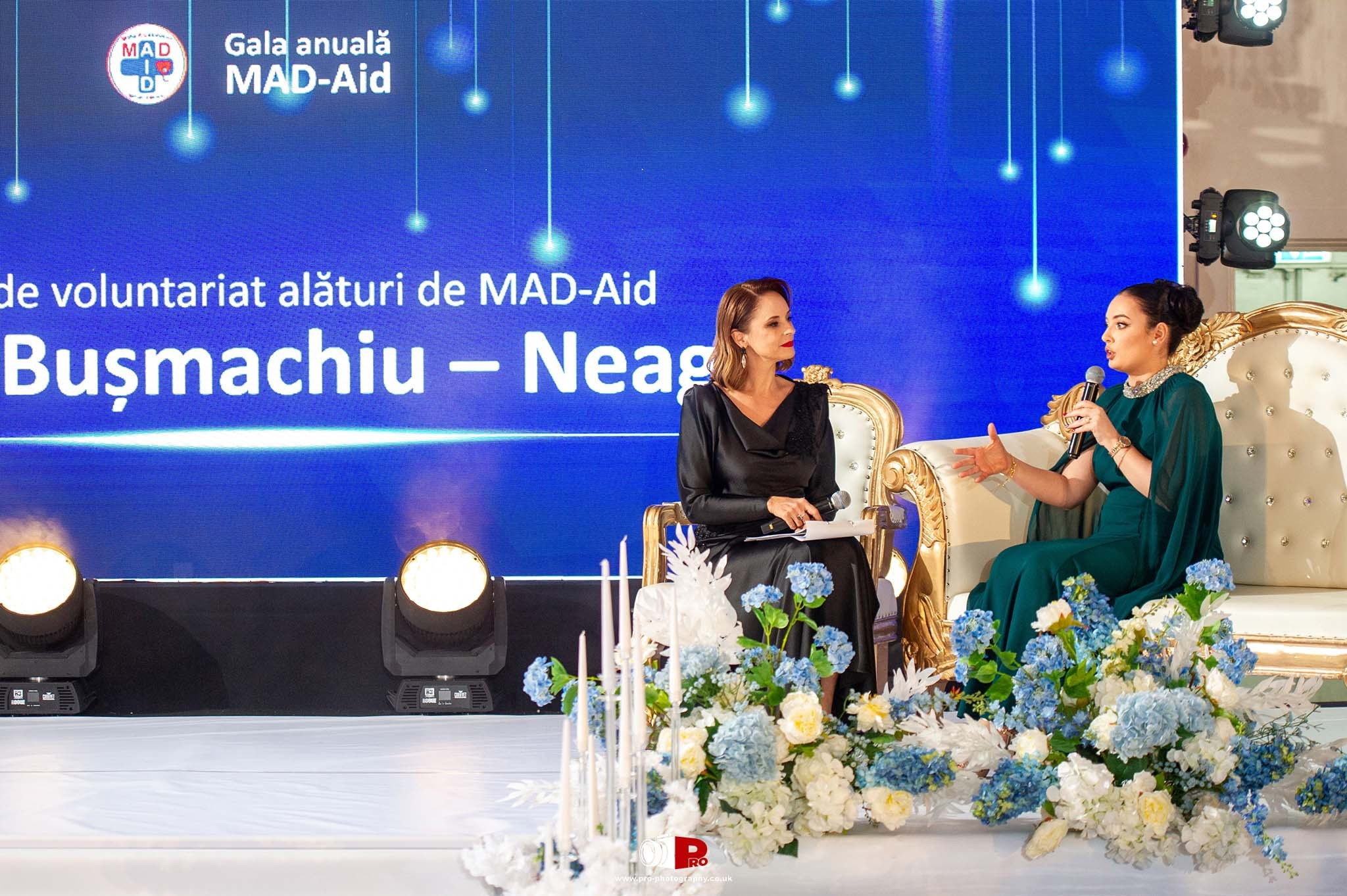 Two elegantly dressed women are having an interview on stage at the MAD-Aid Gala 2024 with floral decor and a blue LED backdrop.