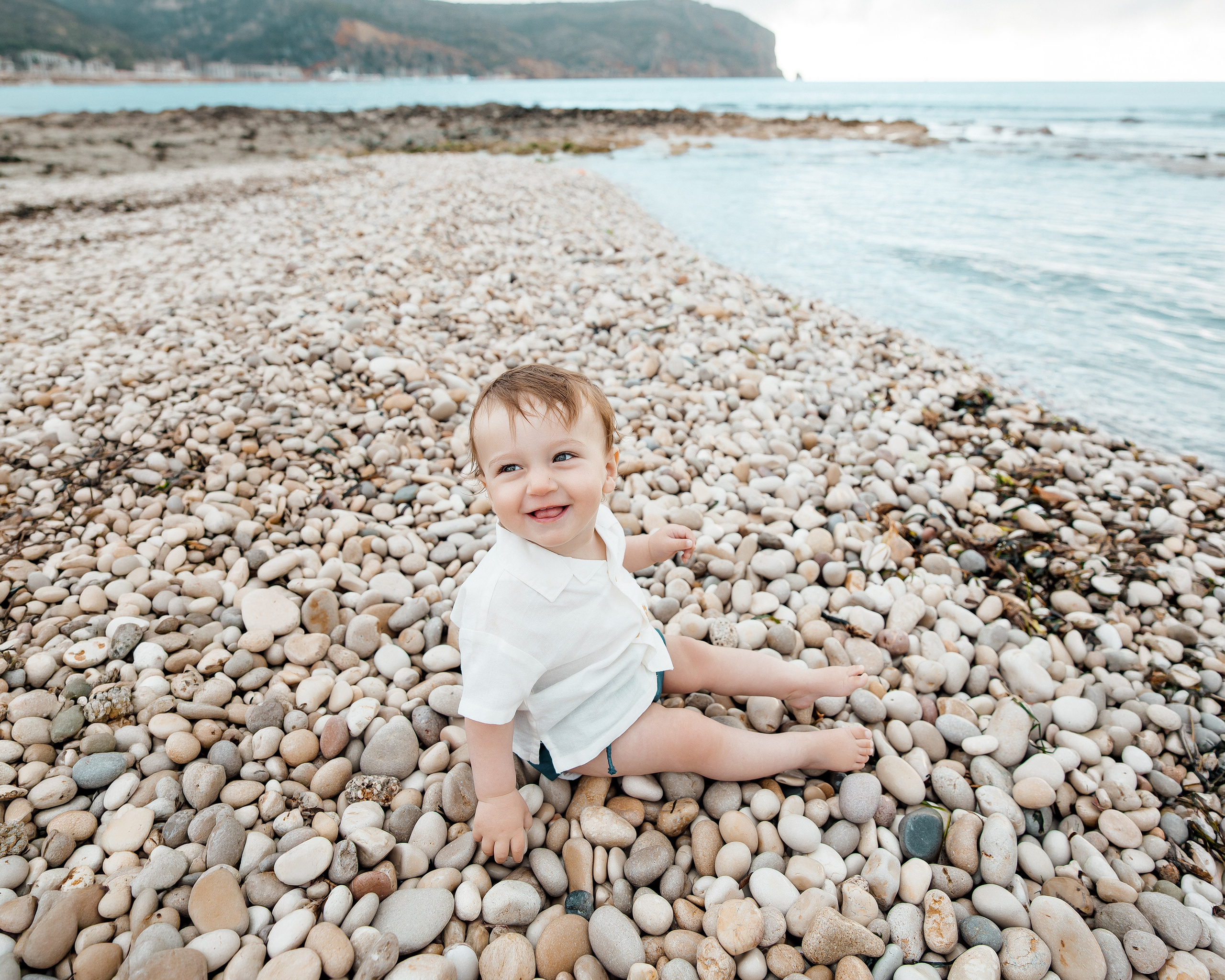 Sesión de fotos familiar en Valencia, España, con un pequeño sentado en una tranquila playa del Mediterráneo cubierta de piedras naturales, disfrutando de un relajante momento junto al mar. Un retrato infantil natural, ideal para fotografía familiar, sesiones de fotos familiares, fotografía infantil, sesiones familiares en la playa, fotografía de vacaciones y fotografía familiar de destino en Valencia y toda España.