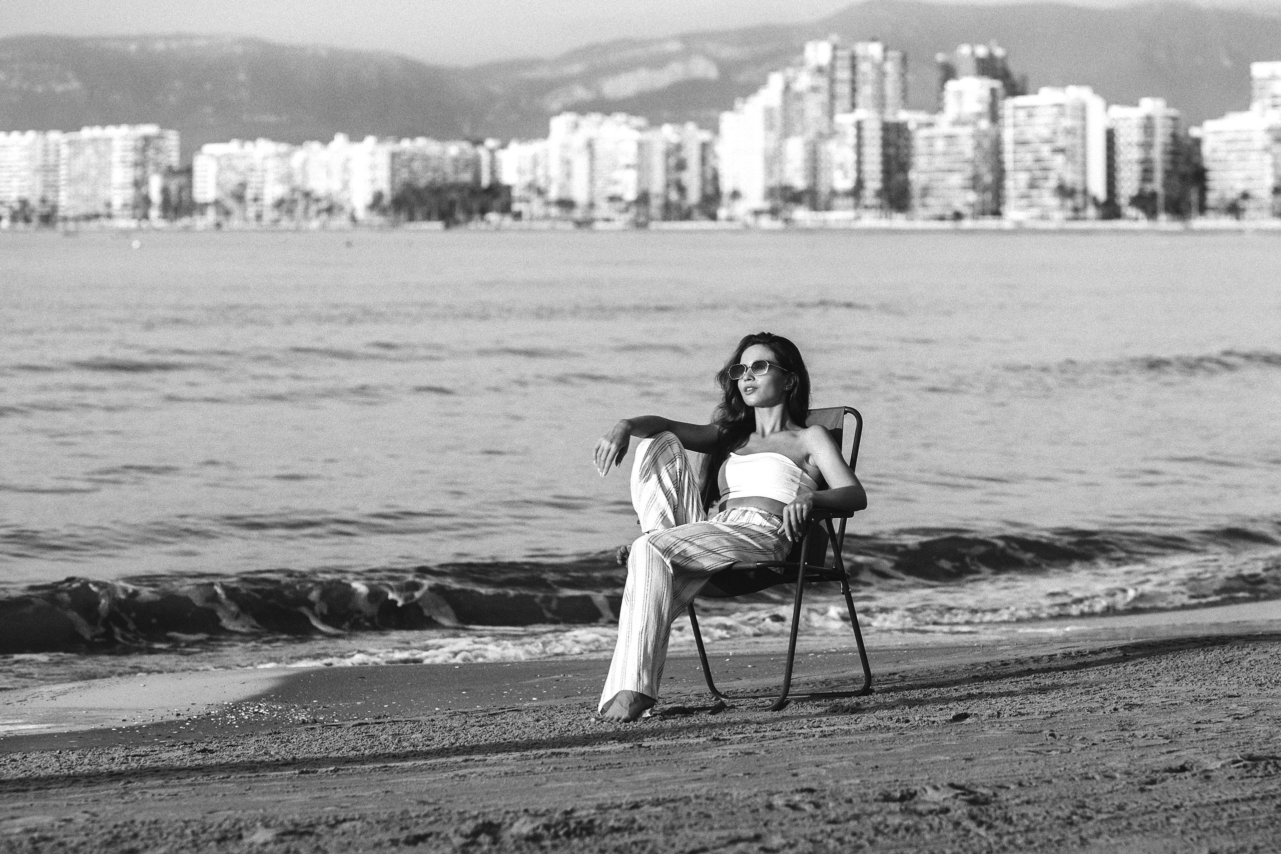 Черно-белый портрет расслабленной женщины в солнцезащитных очках, сидящей на стуле на пляже, с панорамой города Валенсия, Испания, на заднем плане