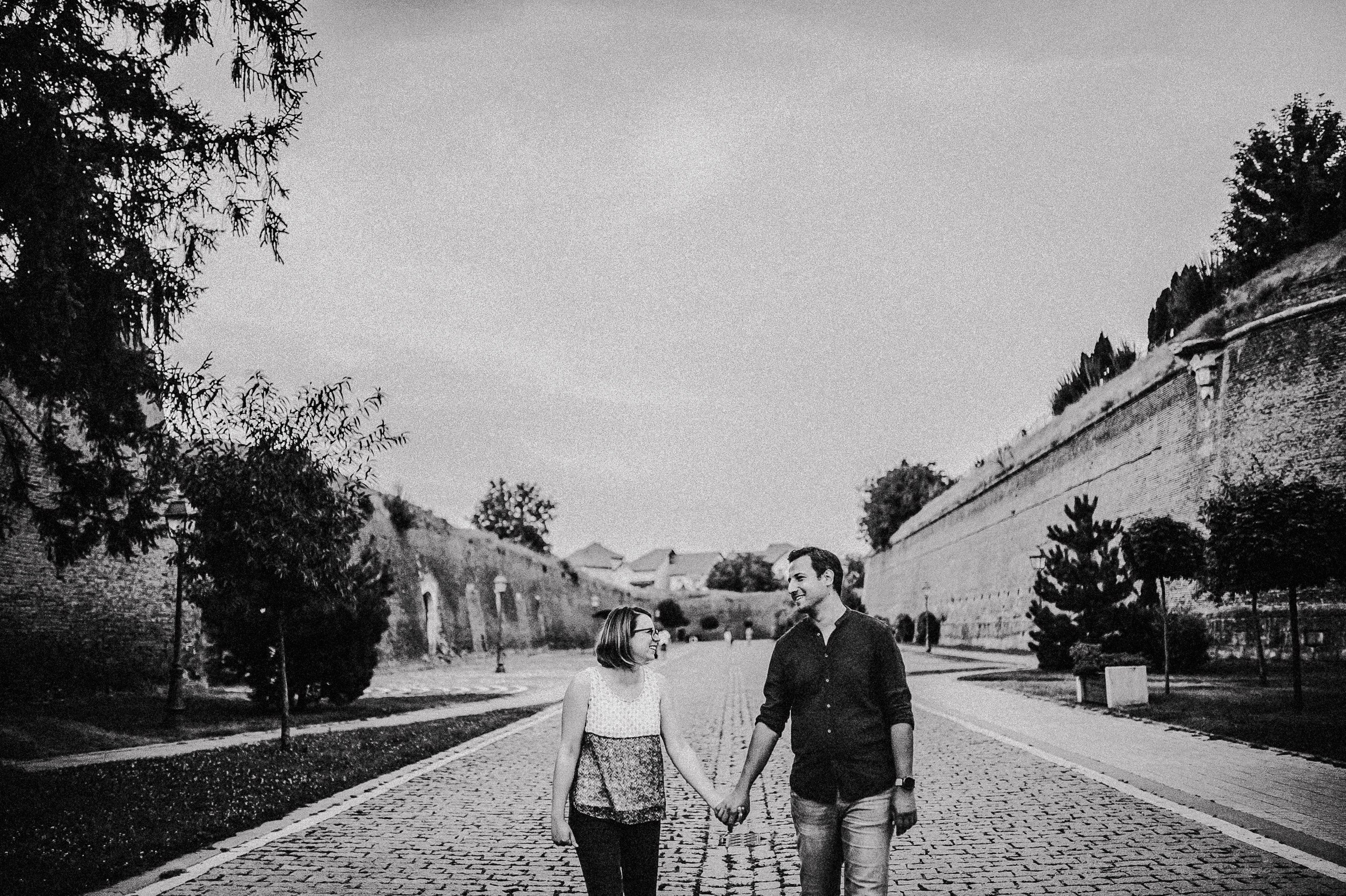 Alina și Ionuț merg de mână pe aleea pietruită dintre zidurile cetății din Alba Iulia, într-o fotografie alb-negru.