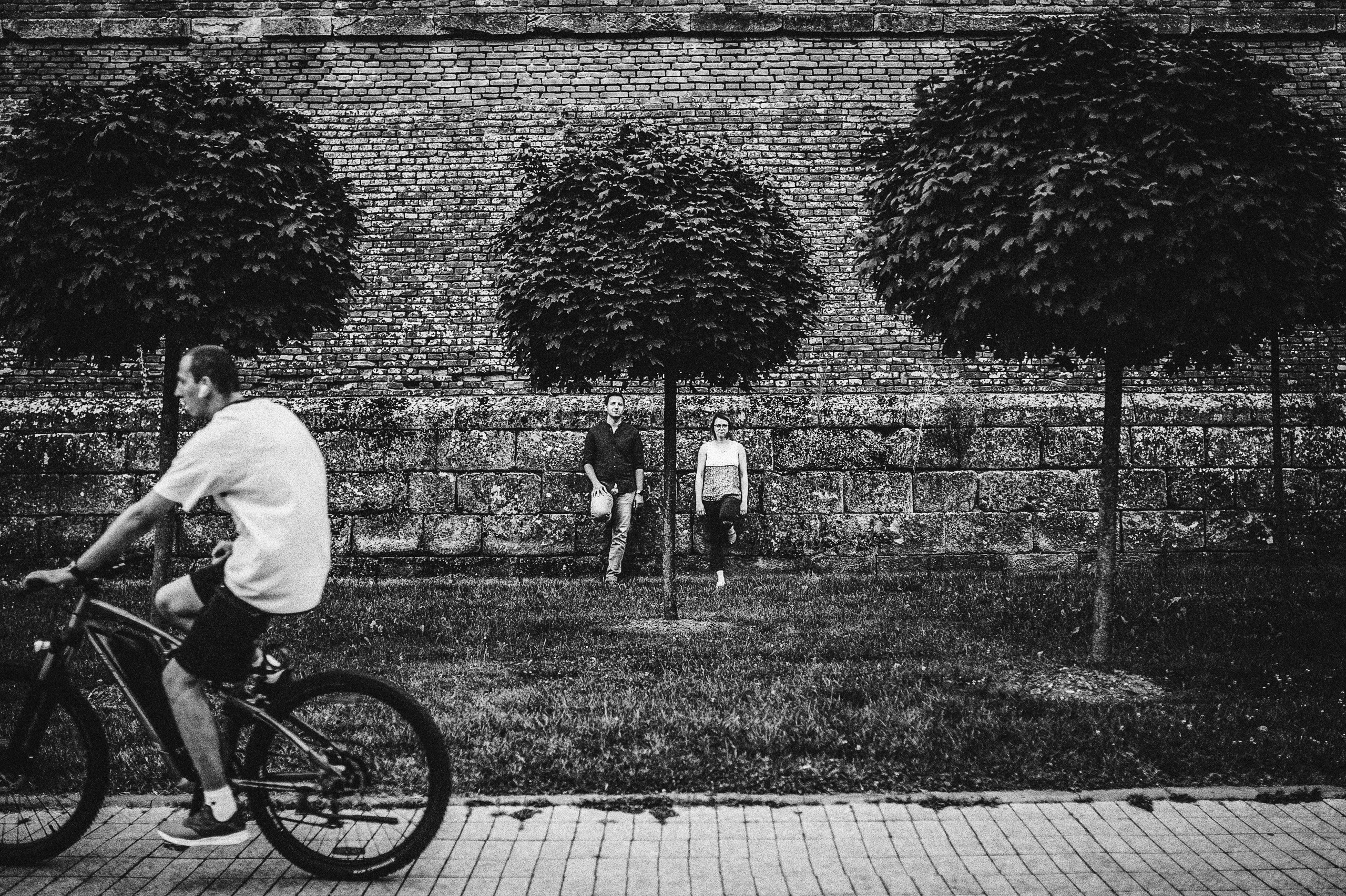 Un biciclist traversează prim-planul, iar Alina și Ionuț stau lângă zidul cetății, într-o fotografie alb-negru.