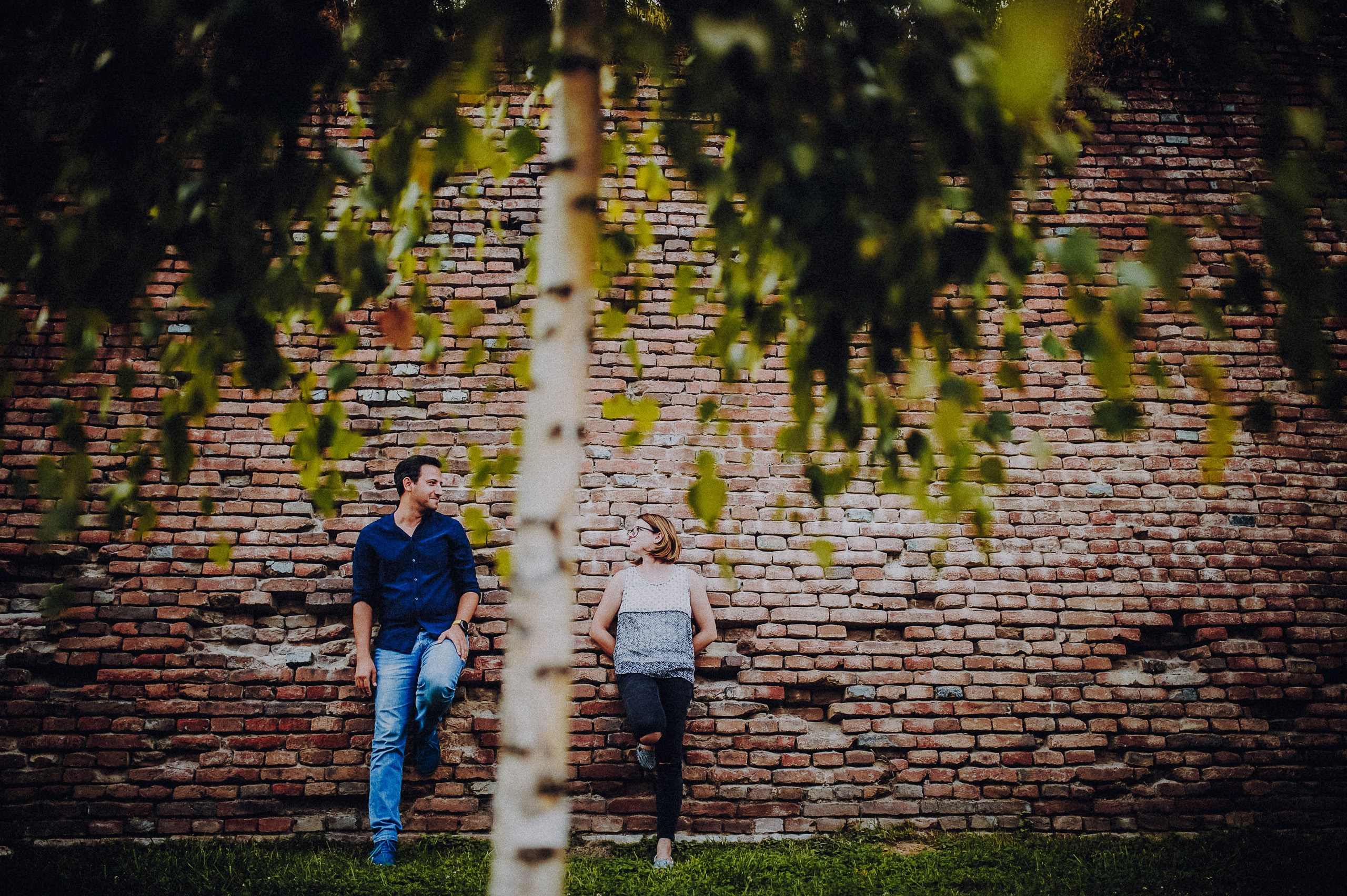Alina și Ionuț își zâmbesc lângă un zid de cărămidă, fotografiați printre frunzele unui copac.