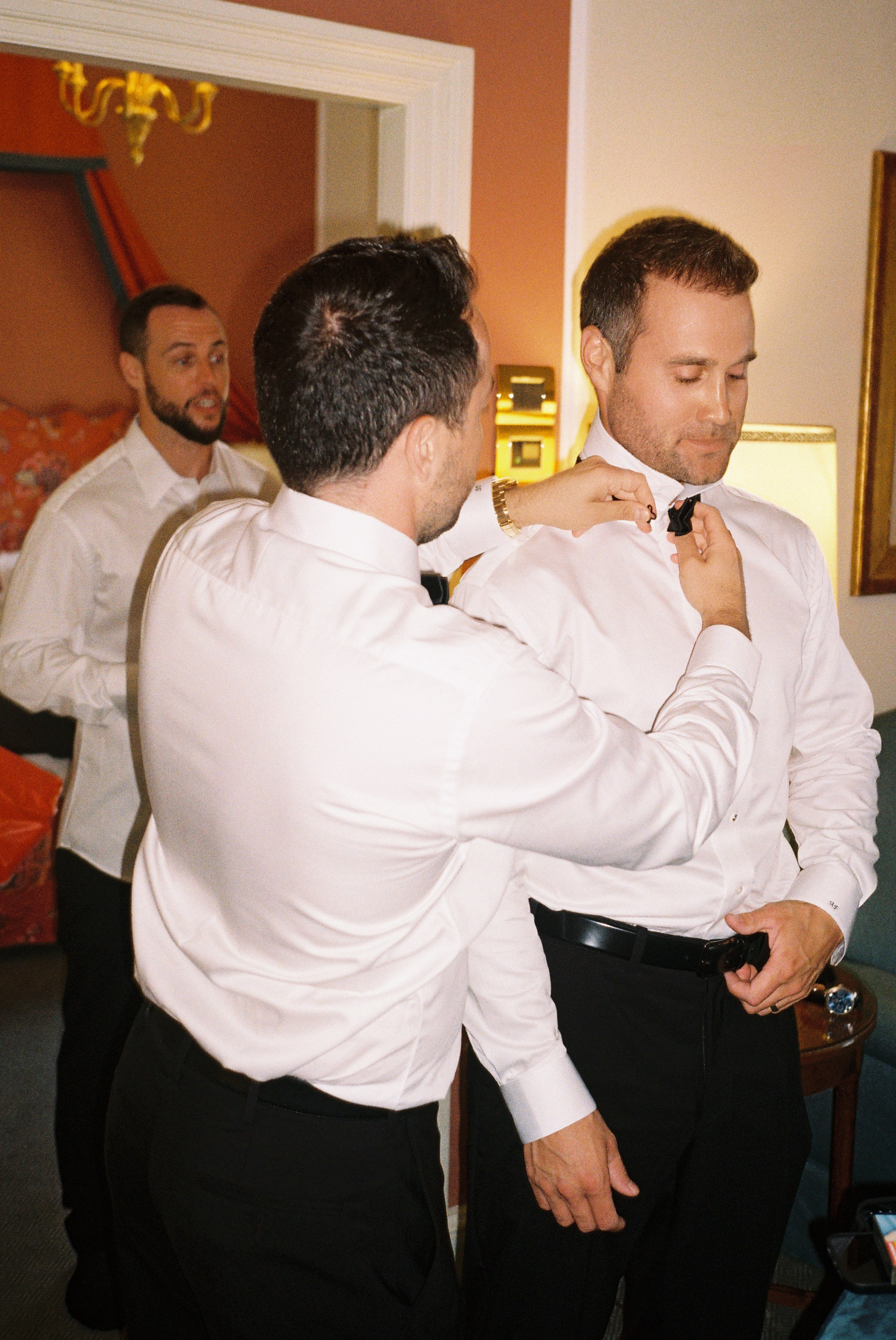 Groomsmen helping each other adjust bow ties, preparing together for the wedding ceremony.