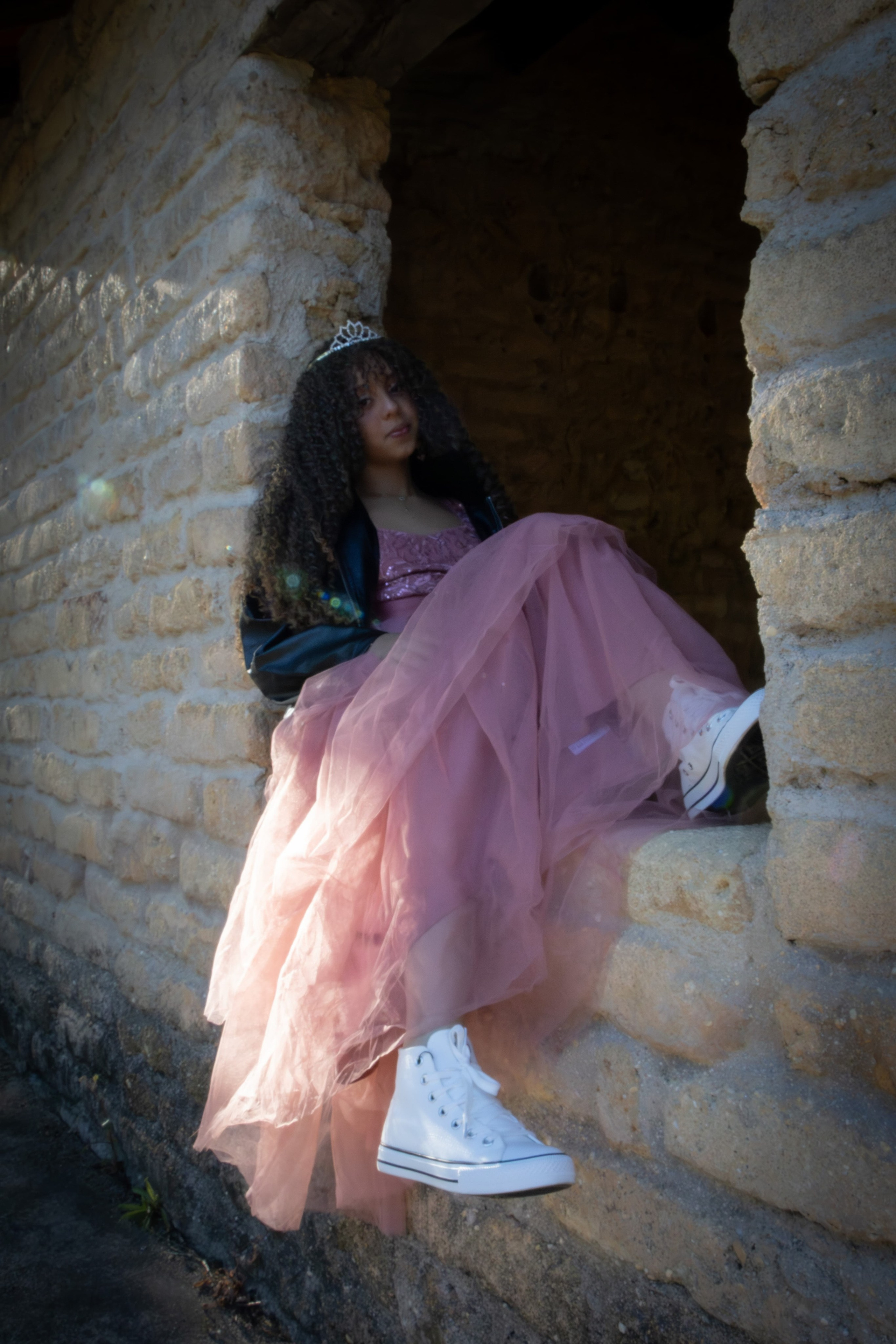 Ensaio feminino fine art. Jovem sentada em uma grande abertura de janela de pedra antiga, usando um vestido rosa longo e volumoso em um ambiente rústico.