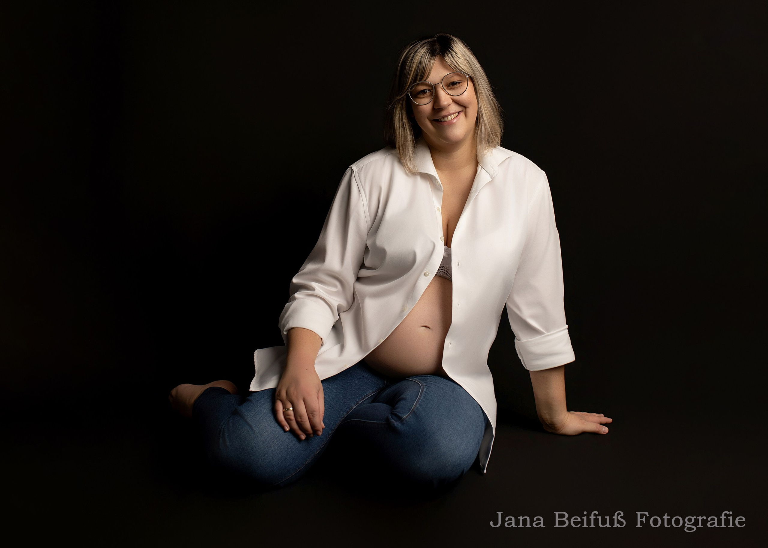 Werdende Mama sitzt auf dem Boden und lächelt schwarzer Hintergrund Fotografin Jana Beifuß in Weißenhorn bei Memmingen Illertissen Ulm