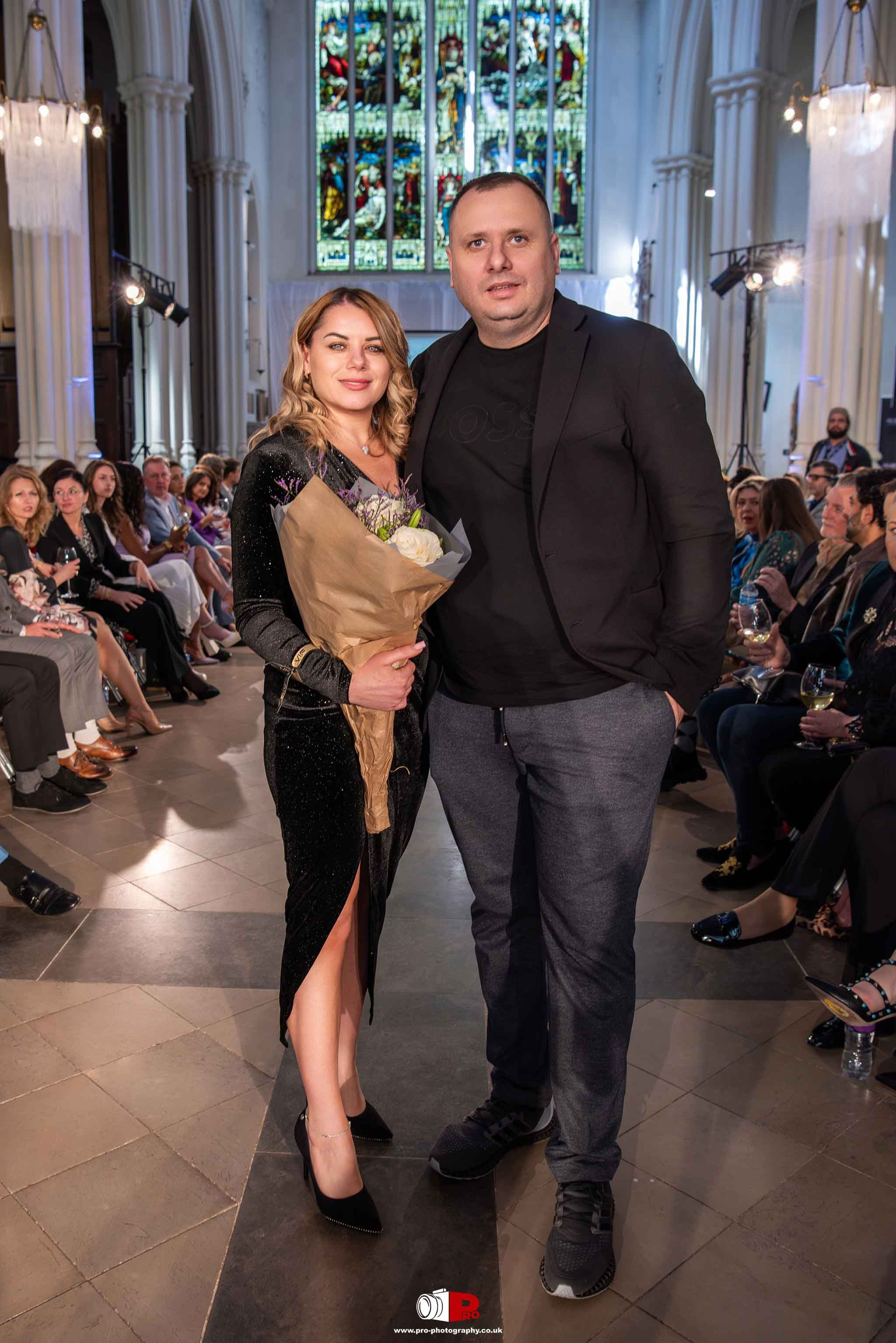 A man and a woman are posing with a bouquet at a prestigious event in a venue with stunning stained-glass windows.
