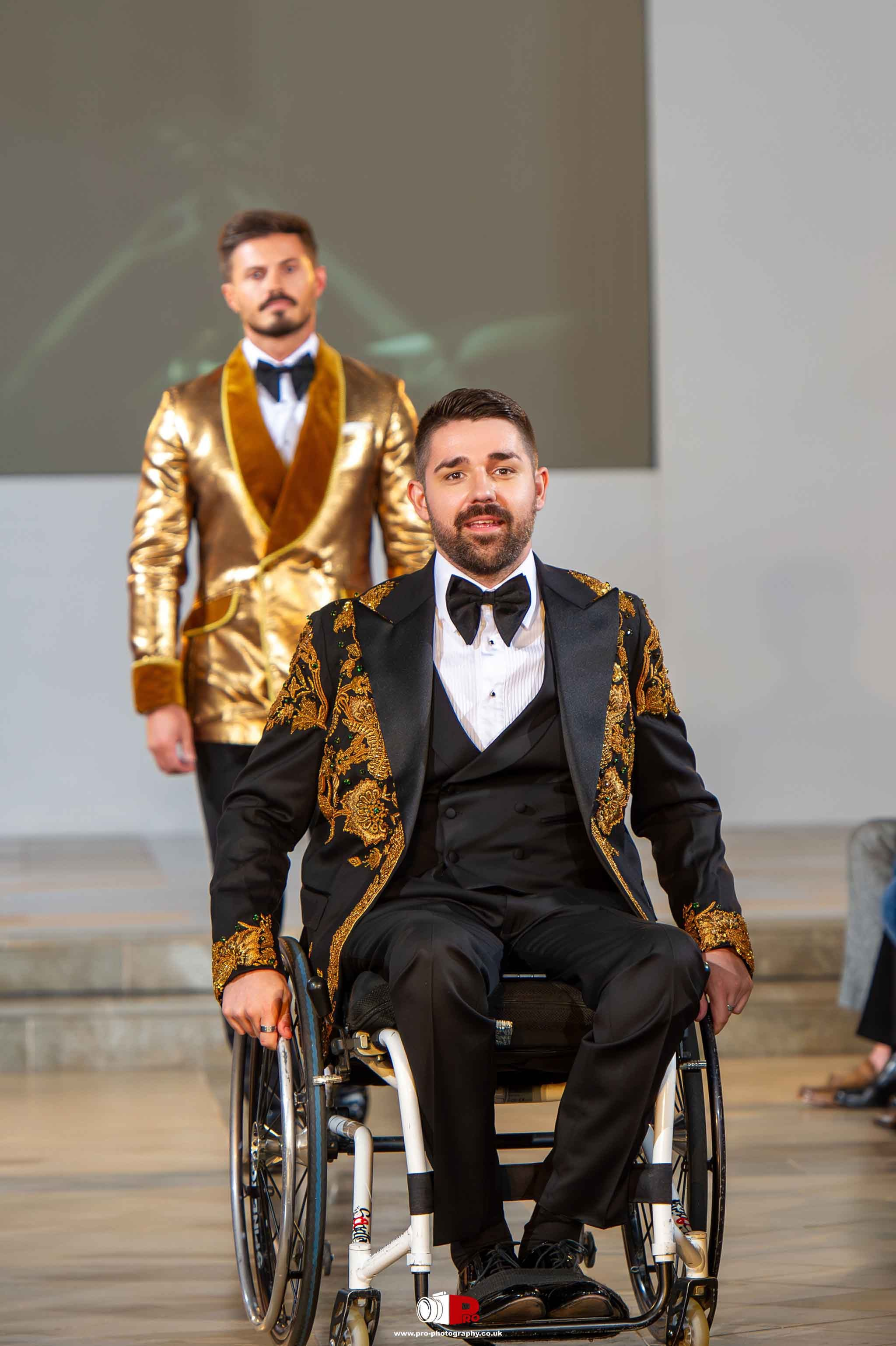 A male model in a wheelchair dressed in a stylish black and gold tuxedo confidently moves down the runway at a fashion event.