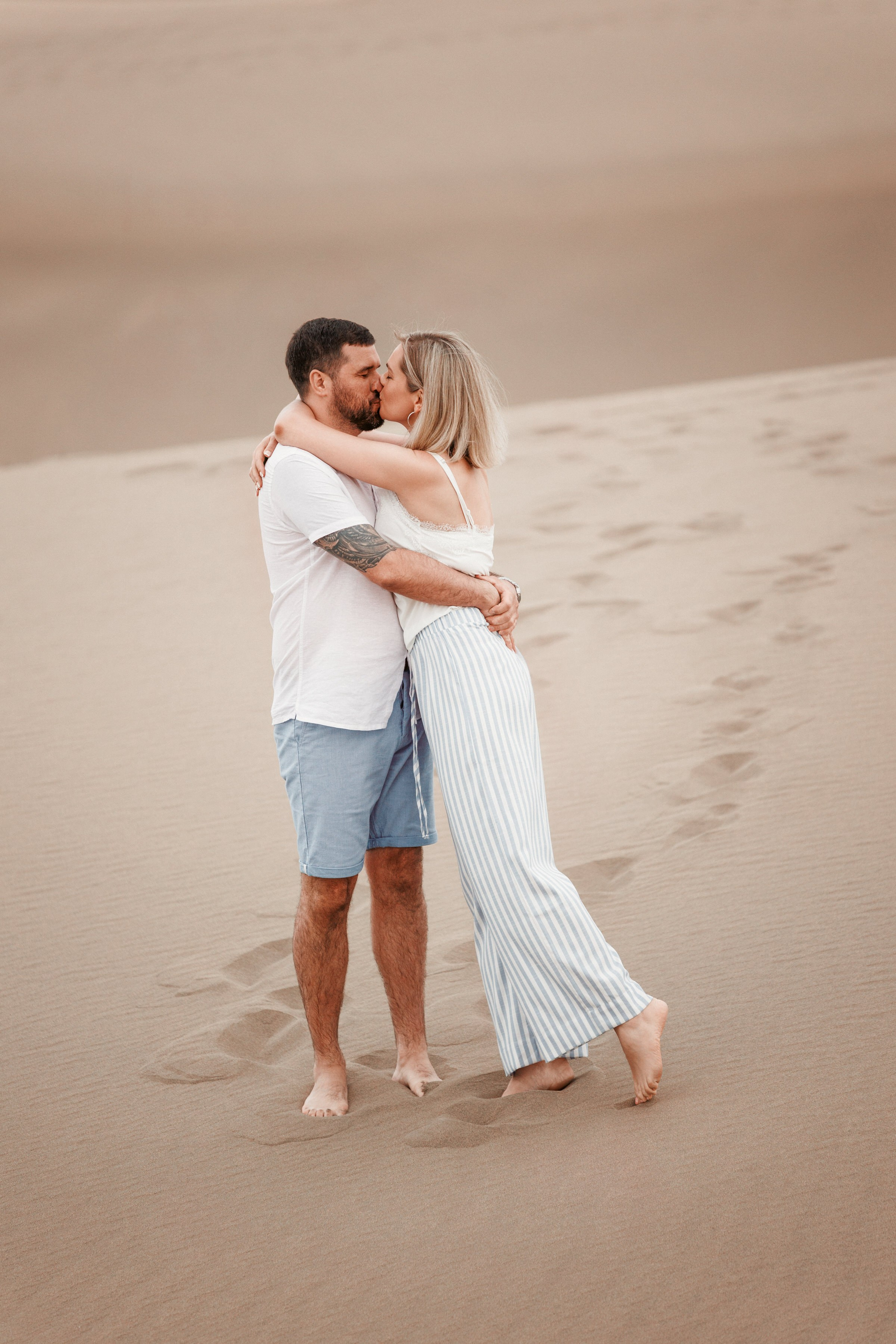 Photographer Gran Canaria Masplaomas - A couple embraces and kisses on a sandy beach, with the woman in a striped dress and the man in shorts and a white shirt.