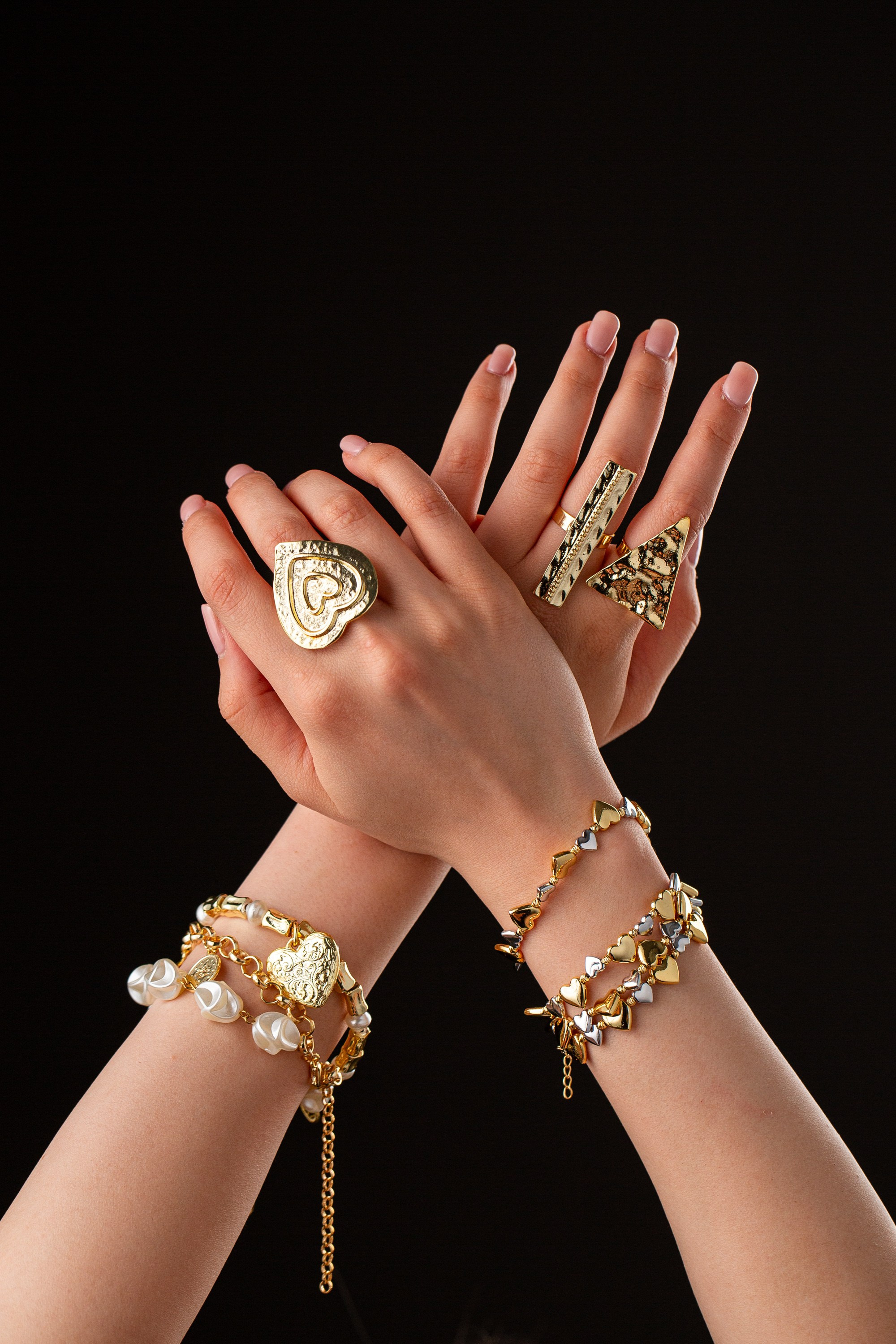 Ürək formalı üzük, üçbucaqlı üzük və ürək və mirvari detallı bilərziklər olan qızıl zinət əşyaları olan əllər.