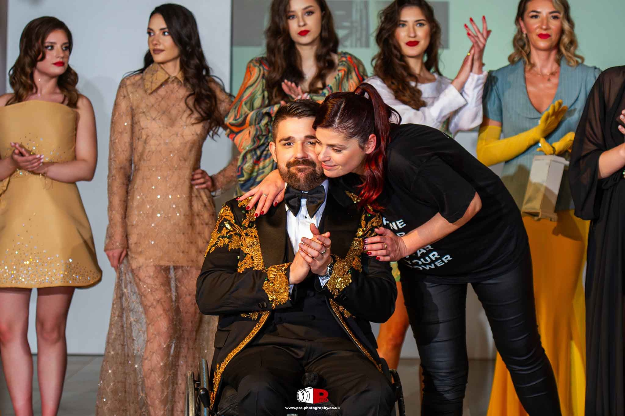 Man in a wheelchair being embraced on stage by Andreea Hasie at a fashion event, surrounded by models.