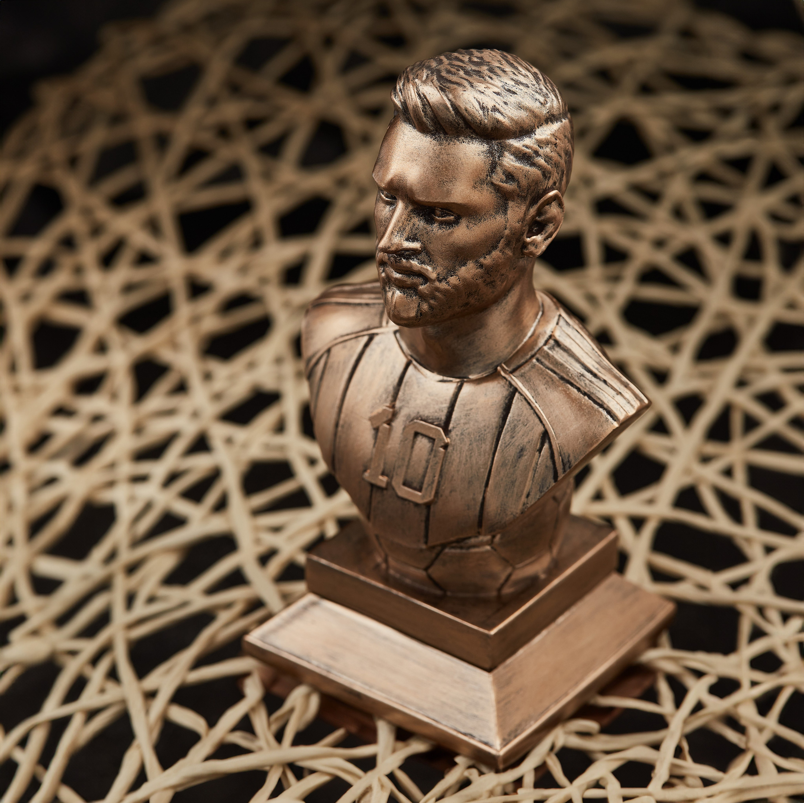 Lionel Messinin 10 nömrəli forması olan bürünc büstü, mürəkkəb toxumalı həsir fon üzərində yaxın plan.