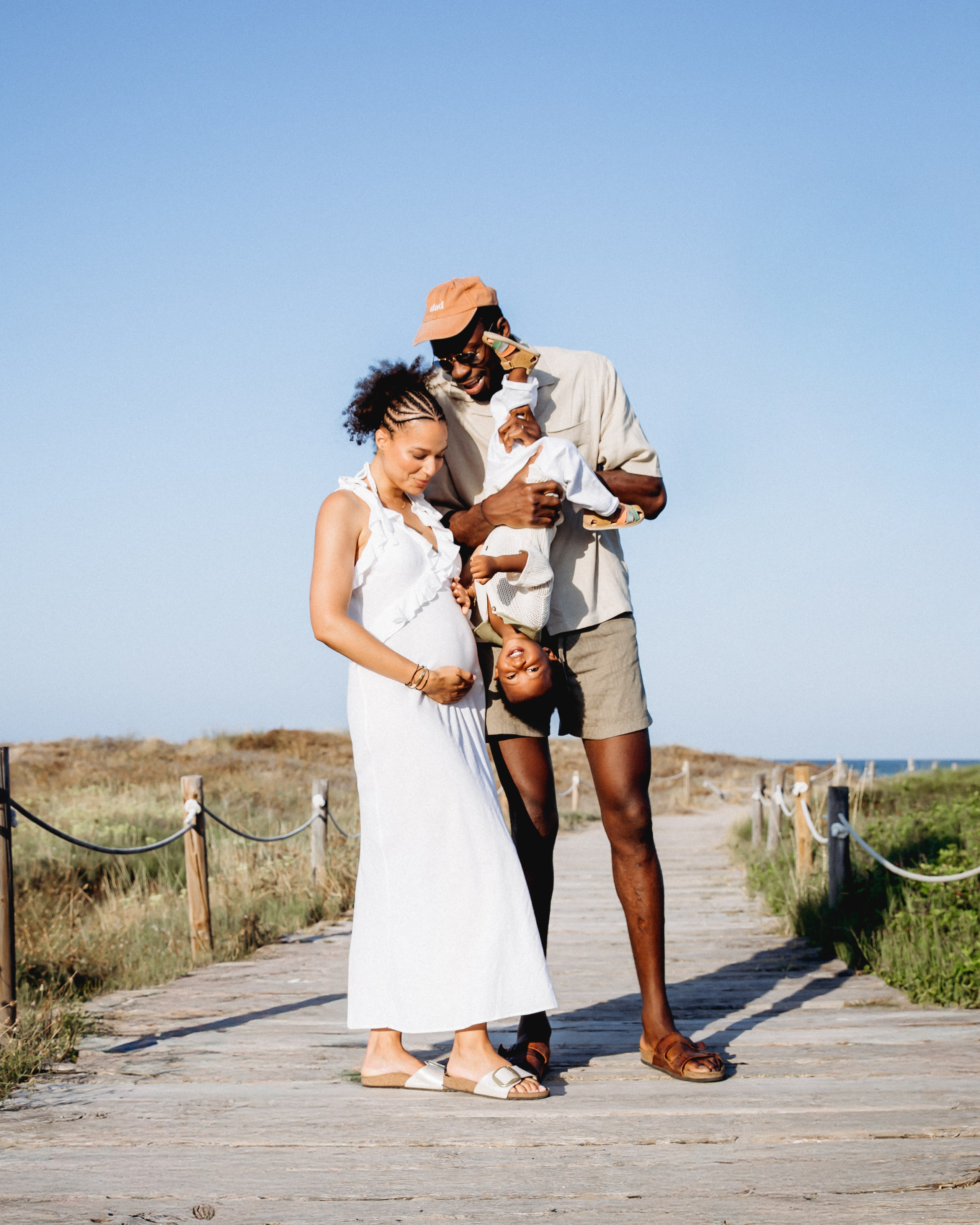 Momento familiar alegre capturado durante una sesión de fotos al atardecer en la playa de Cullera, España — padre jugando con su hijo cabeza abajo, madre embarazada sonriendo. Ideal para quienes desean sesiones fotográficas familiares naturales y emotivas en Cullera y en toda España.