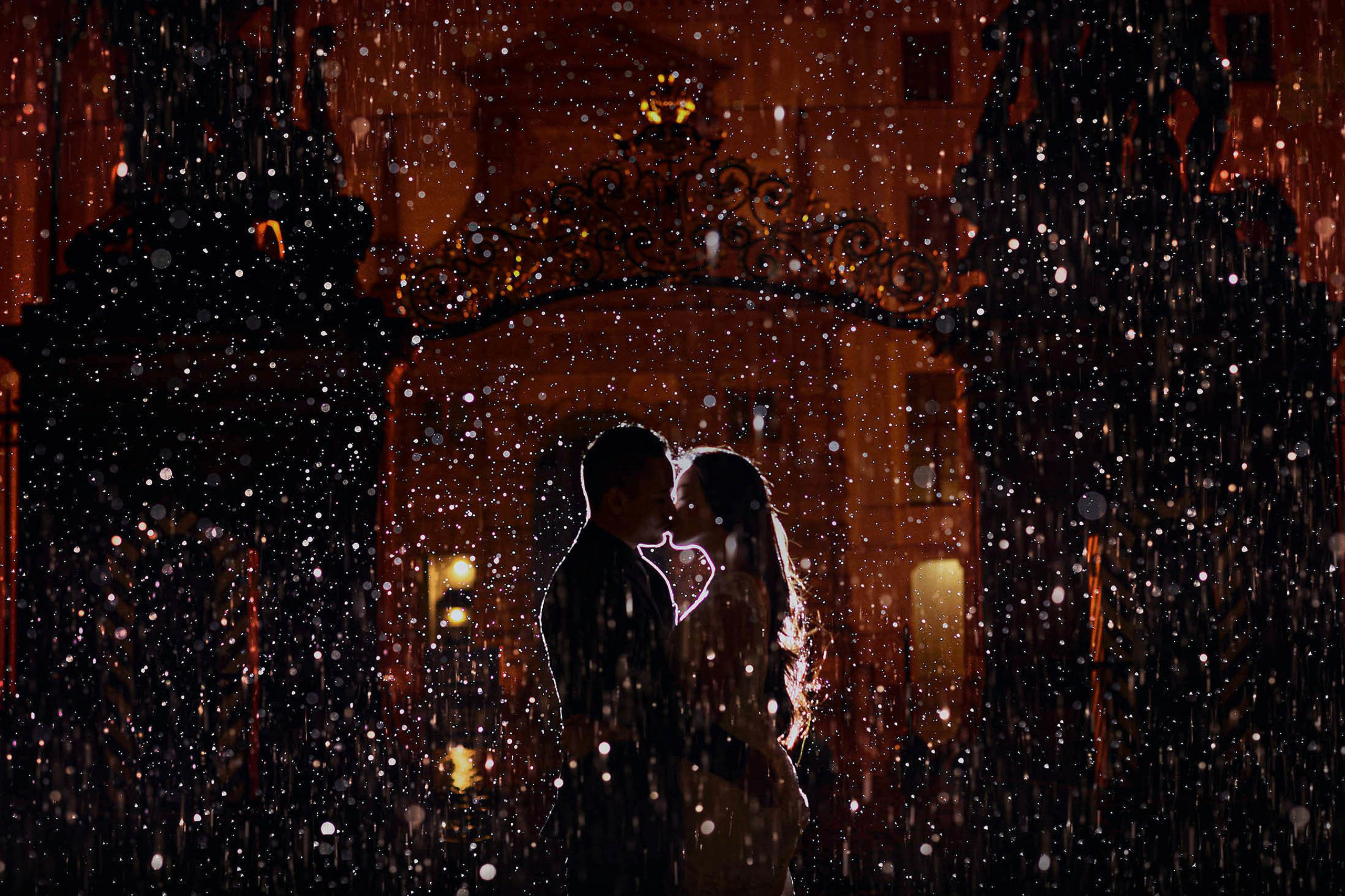 A man and woman are silhouetted against the night time scenery of Prague Castle as rain falls around them