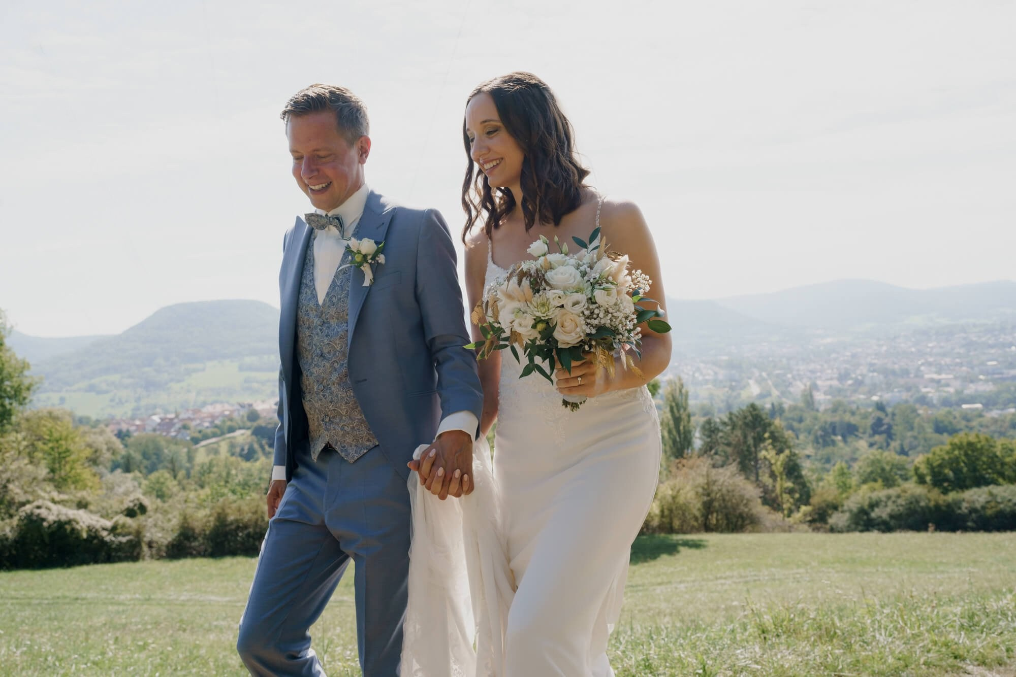 Bride holding a white bouquet and groom walking together laughing, rolling hills of Stuttgart visible behind them