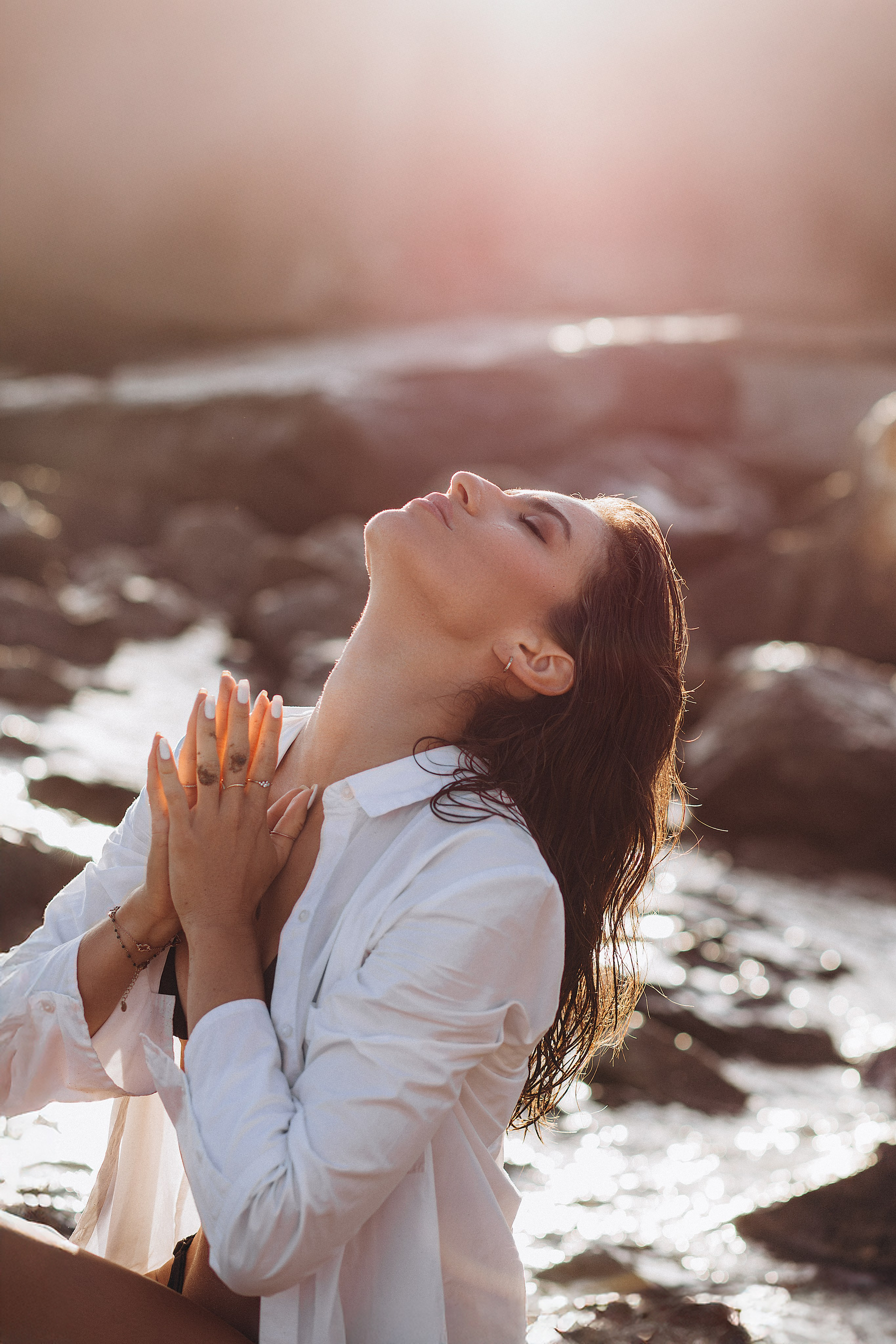 Эмоциональный портрет на пляже в Бильбао, Испания — женщина в белой рубашке с руками у груди и поднятым вверх лицом в золотом свете заката. Пример выразительной и душевной портретной съёмки для тех, кто ищет профессиональные фотосессии в Бильбао и на севере Испании.