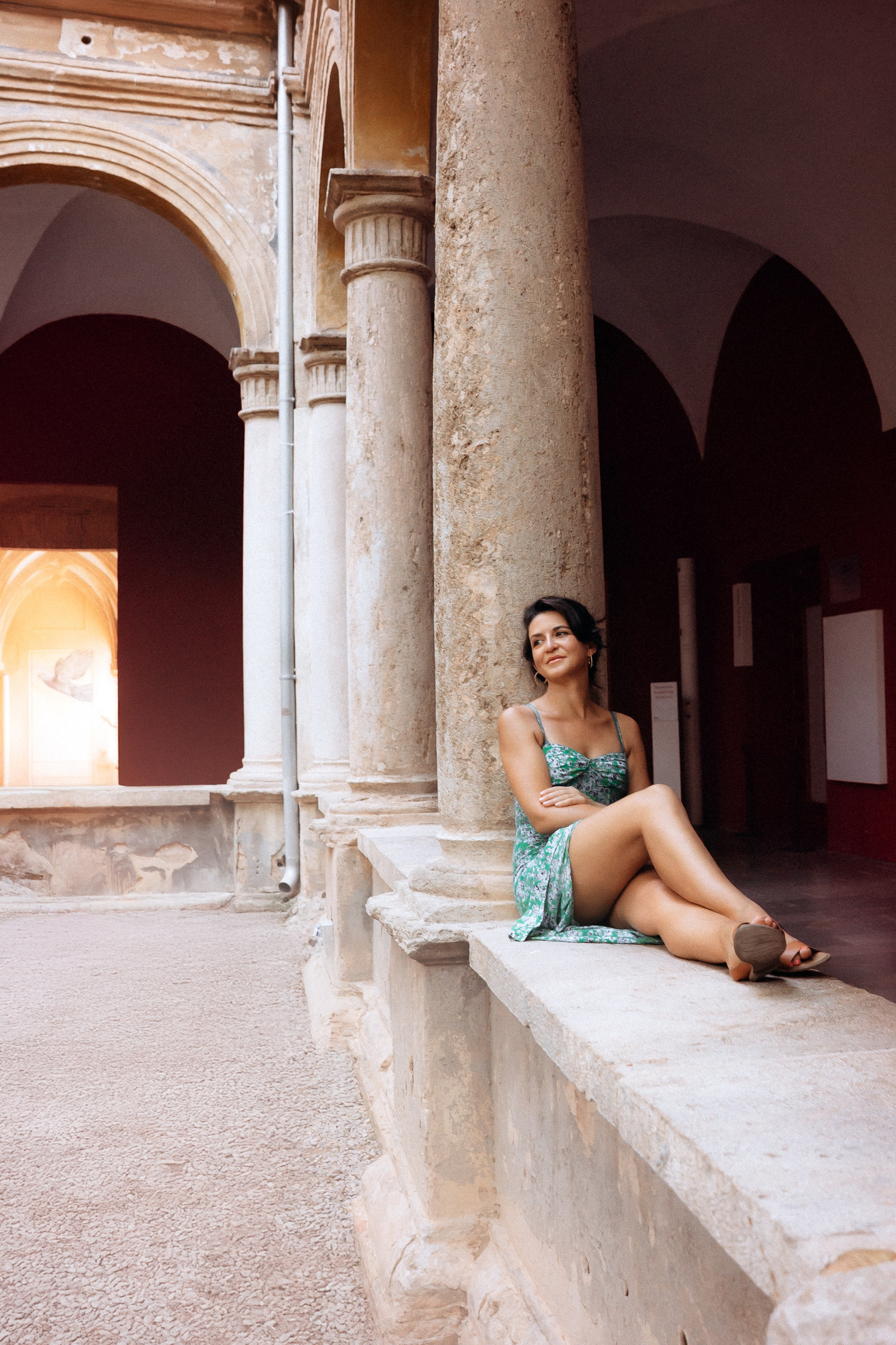 Женская фотосессия в Валенсии, Испания, со стильной женщиной, сидящей на каменном выступе между историческими арками и колоннами, снятая в тёплом естественном свете с элегантной, вневременной атмосферой — идеально для женских фотосессий в Валенсии, лайфстайл- и тревел-портретов, художественных женских портретов и профессиональной женской фотографии по всей Испании.