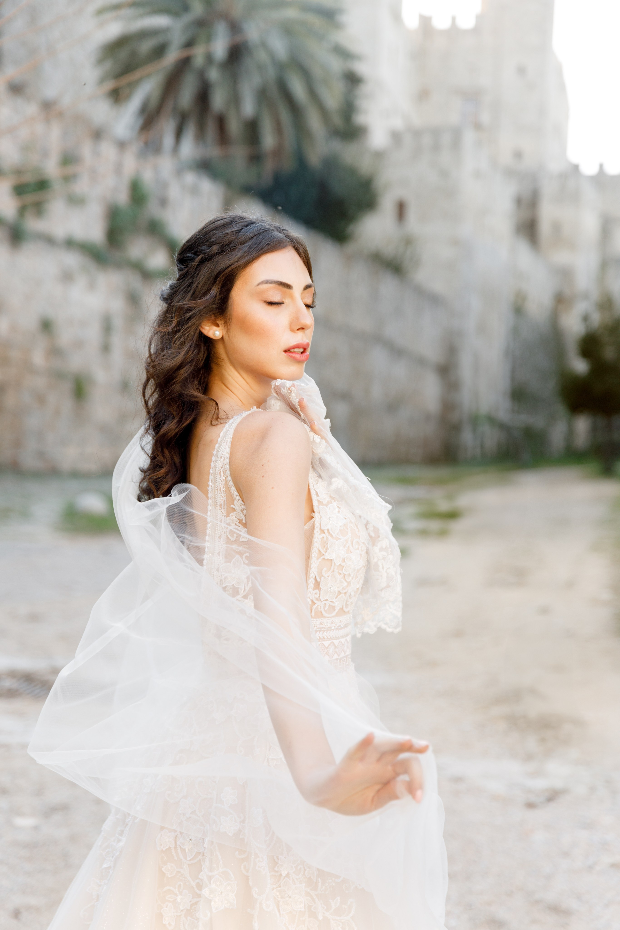 A stunning bride gazes thoughtfully in the enchanting alleys of Rhodes' Old Town, her flowing wedding dress complementing the rustic charm of the cobblestone streets and ancient architecture. The editorial-style portrait captures her poise and the romantic atmosphere of the medieval surroundings, bathed in warm, golden light.