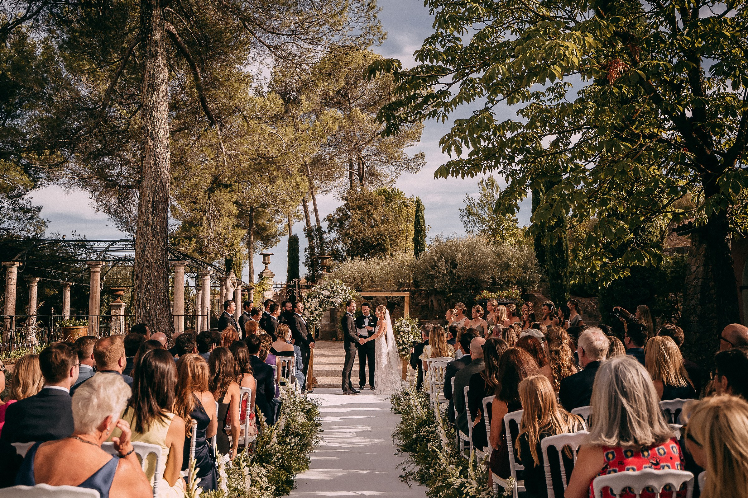 A breathtaking outdoor wedding ceremony in Provence, France, surrounded by lush greenery and tall trees. The bride and groom stand at the altar under a wooden arch adorned with white flowers, while guests sit on white chairs along an aisle lined with floral arrangements, all illuminated by soft sunlight filtering through the trees.