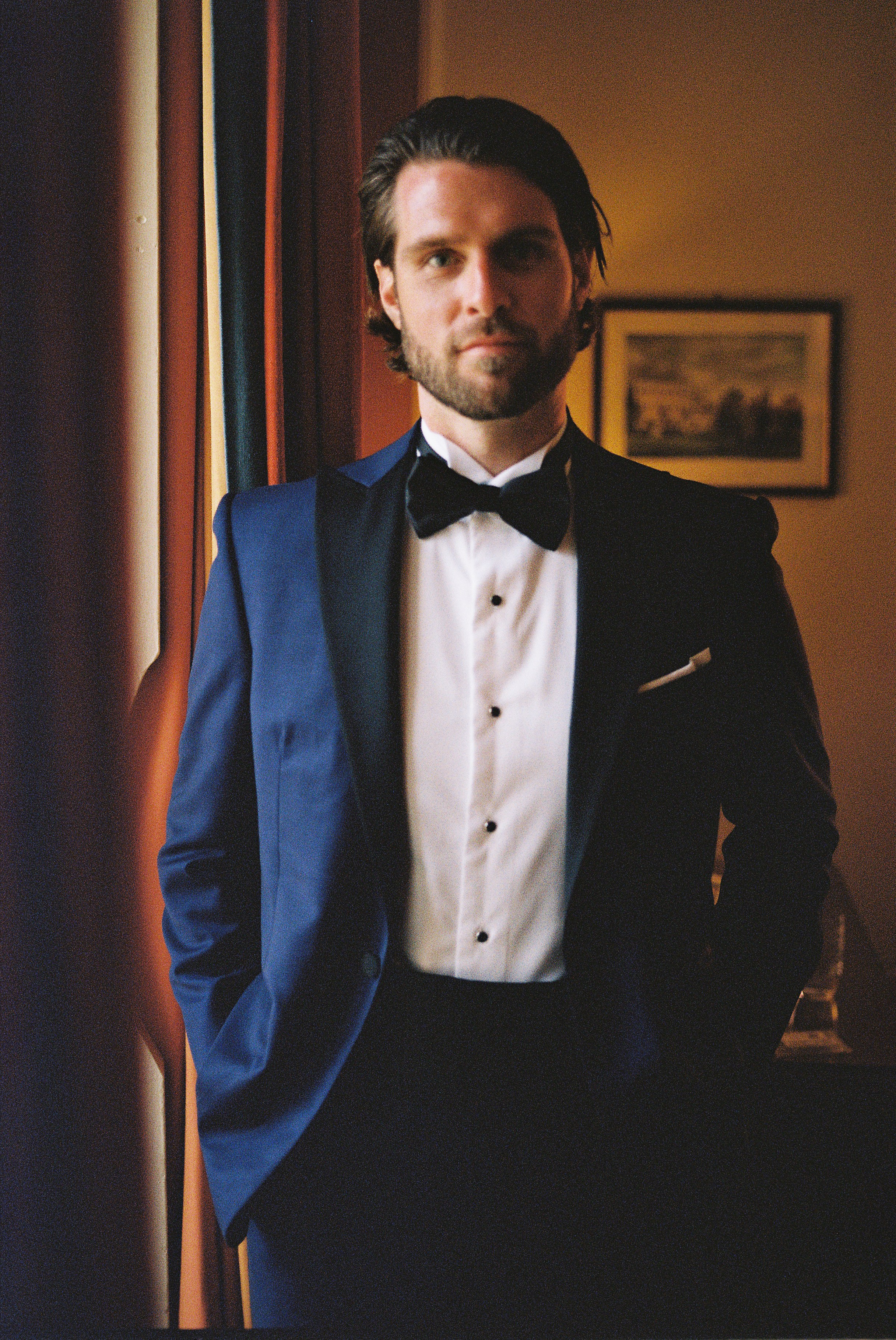 Groom gazes thoughtfully, wearing a crisp tuxedo, framed by warm indoor light and vintage decor.