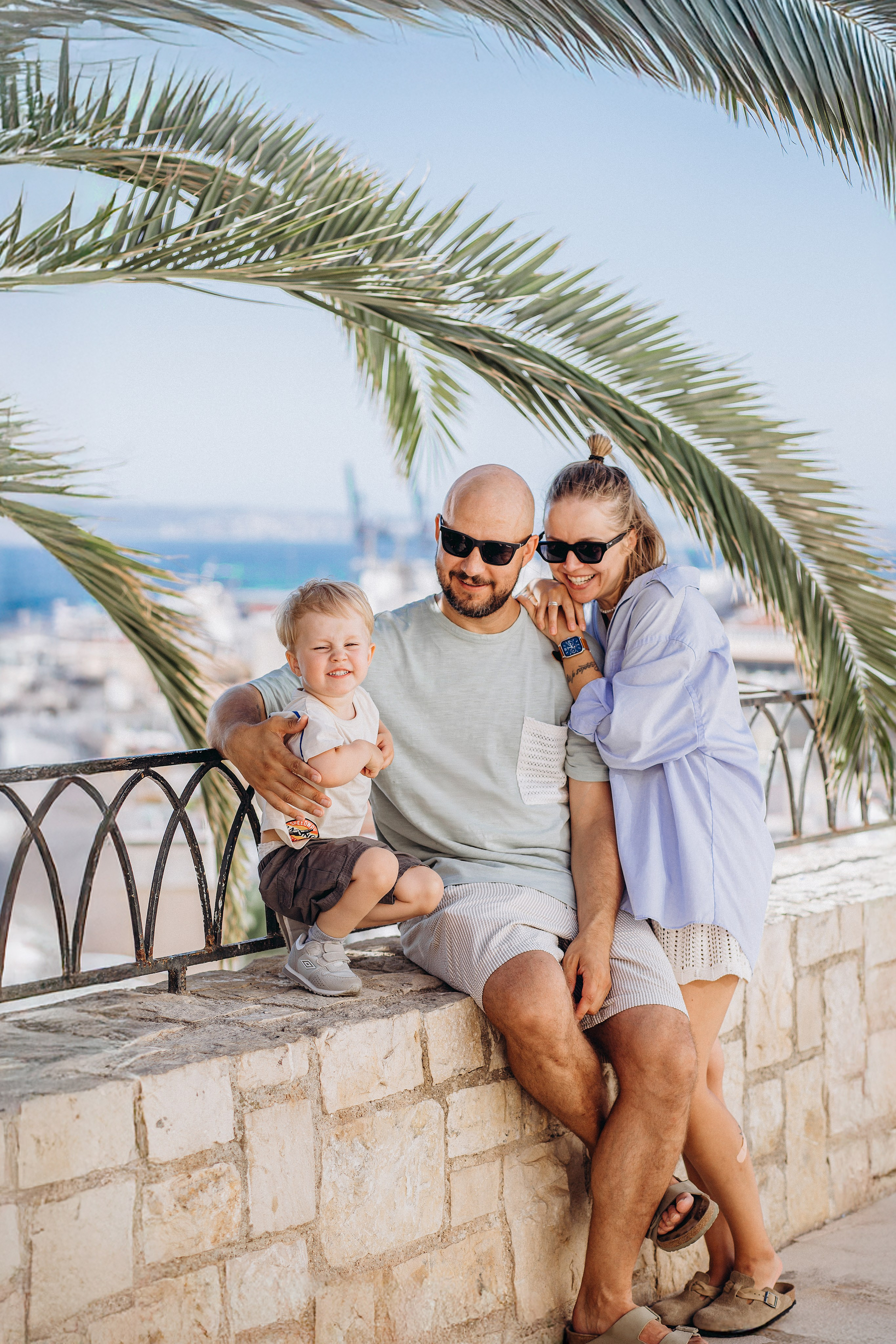 Familia feliz sentada en una terraza de piedra con palmeras y vistas al mar en Alicante, España, durante una sesión familiar luminosa y alegre. Ejemplo perfecto de fotografía vacacional relajada para familias que buscan sesiones profesionales en Alicante y en toda España.
