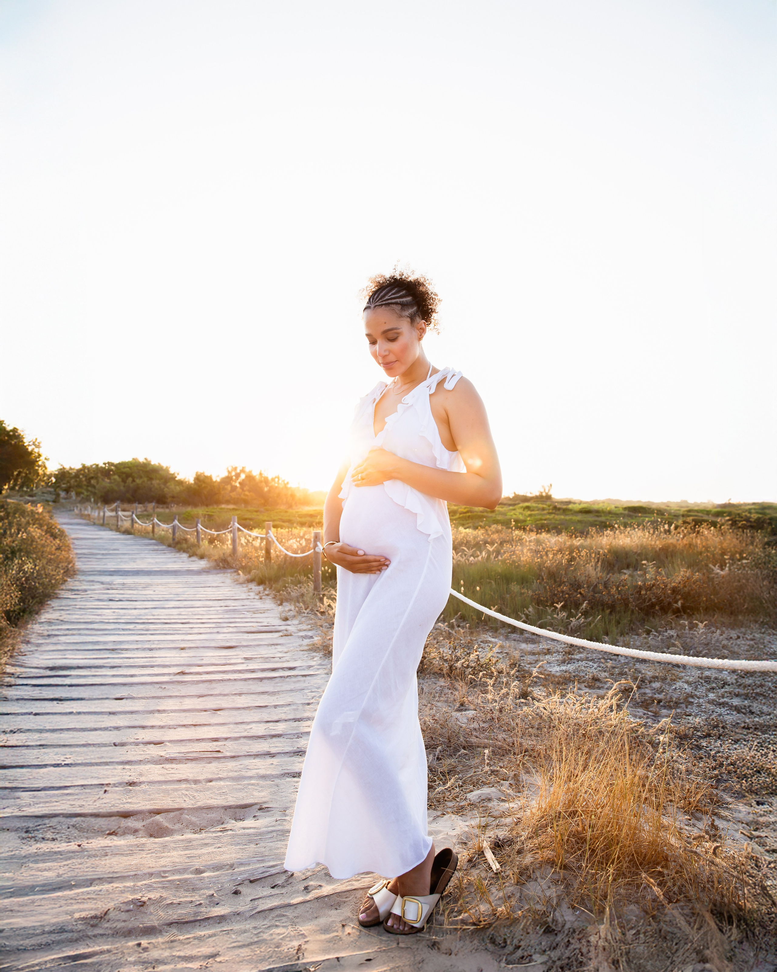 Sesión de embarazo al atardecer en Alicante, España — mujer embarazada caminando sobre una pasarela de madera rodeada de hierba dorada, capturando un momento sereno y natural al aire libre en la Costa Blanca.