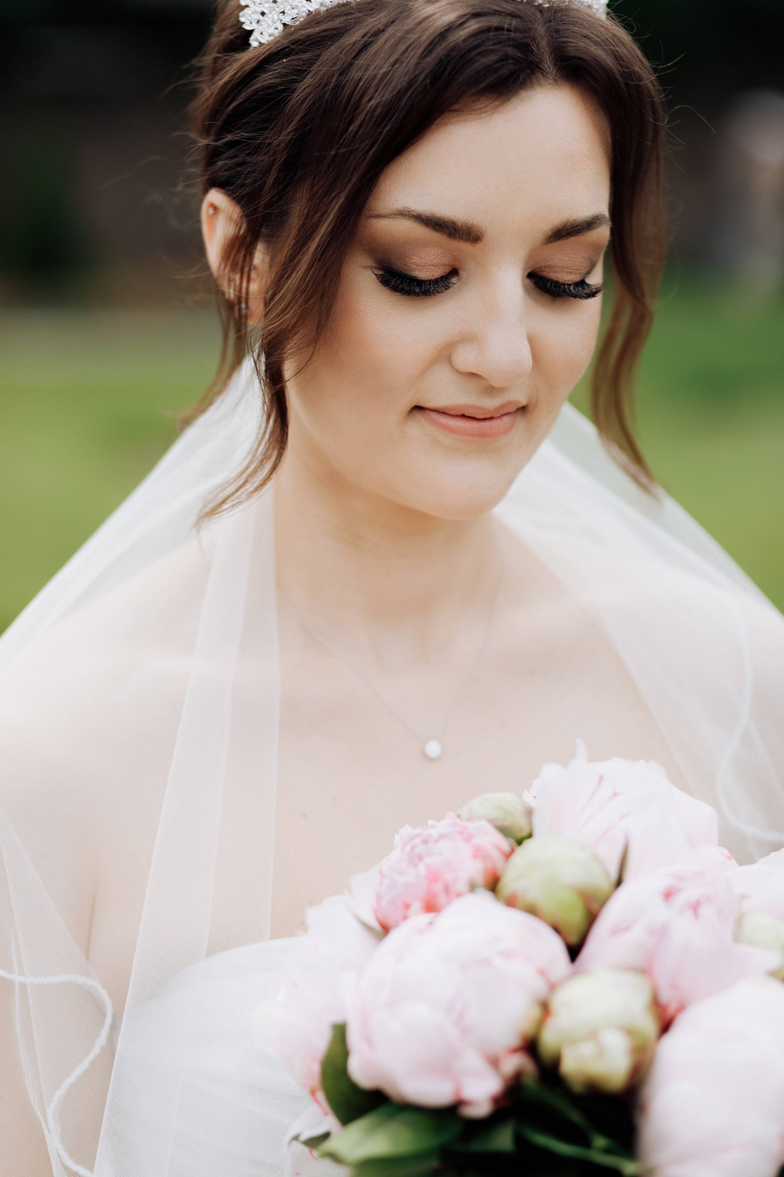 Die Braut schaut auf ihren Brautstrauß mit Pfingstrosen, während ihr makelloser Make-up sichtbar ist. 