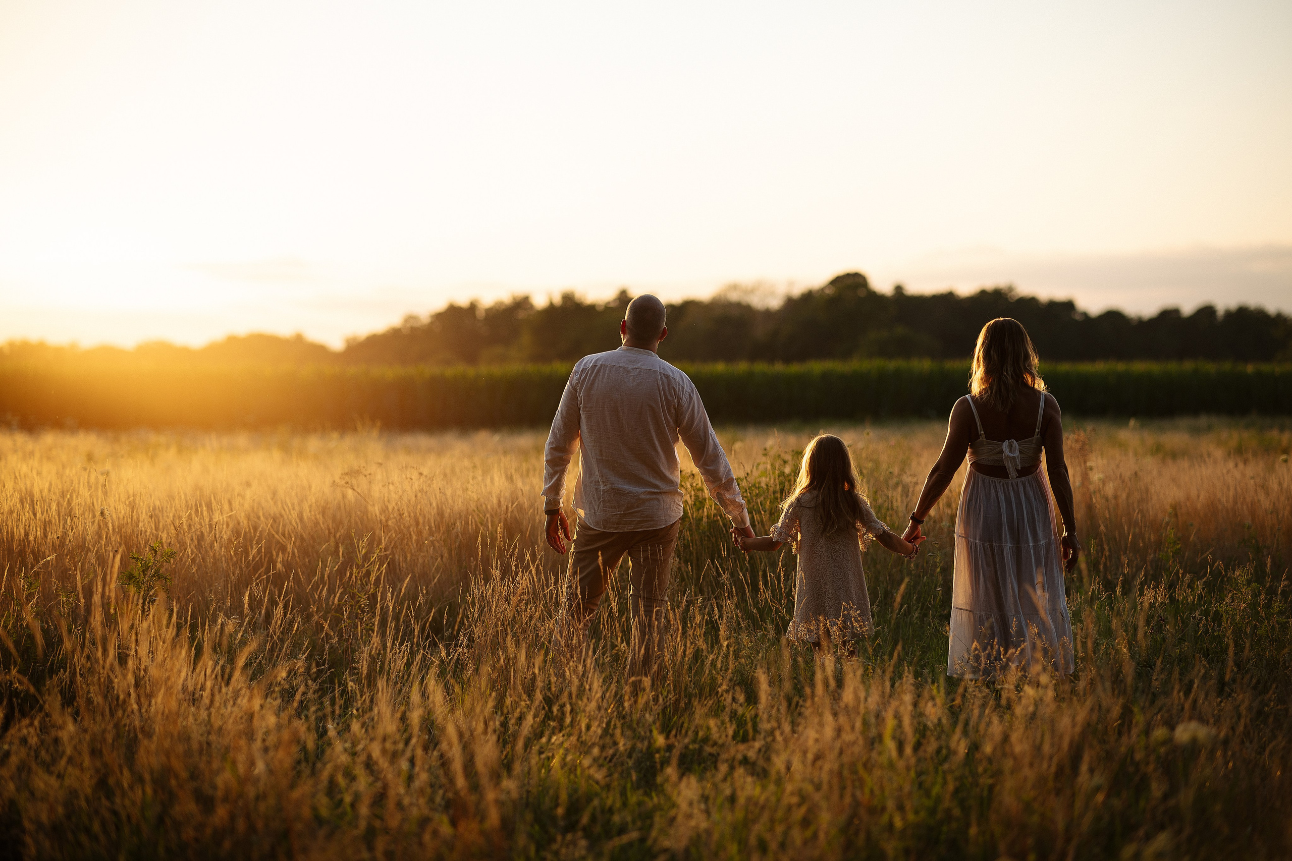 Familie mit schwangerer Mutter, Vater und Tochter geht bei Sonnenuntergang durch ein Feld und blickt in die Abendsonne.