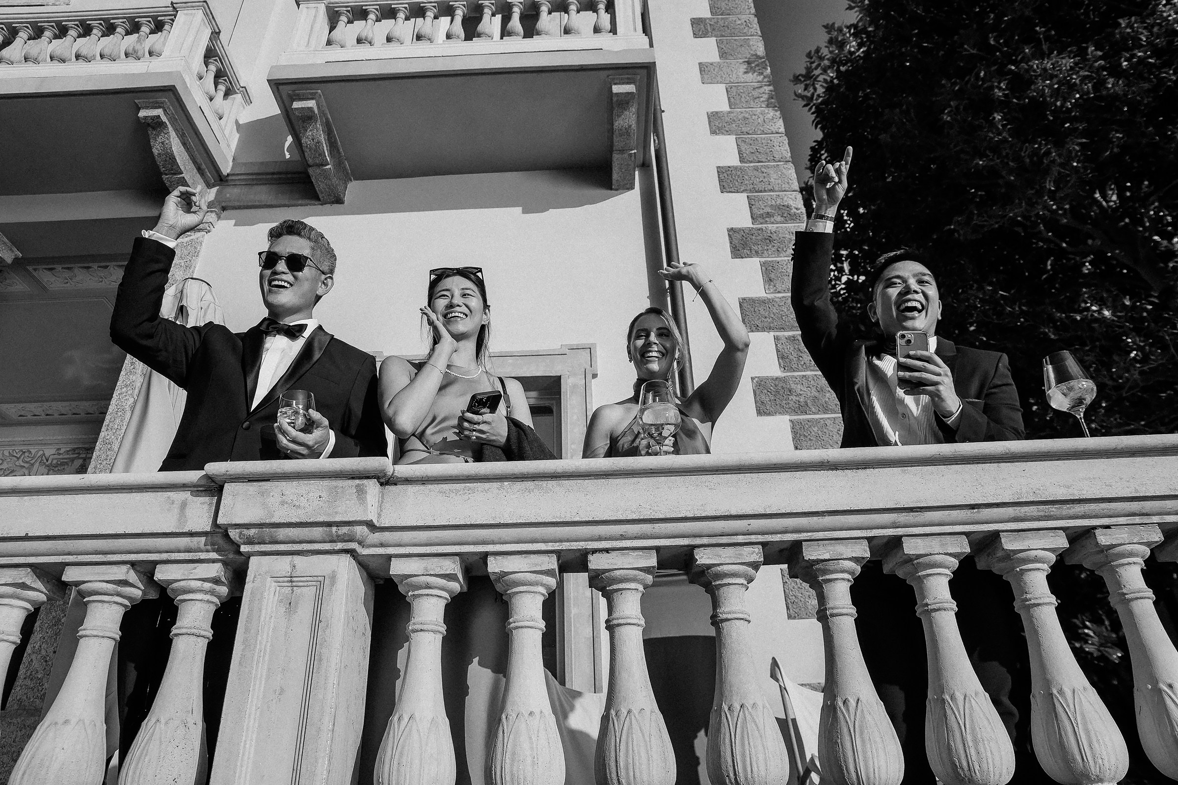 Four elegantly dressed guests standing on a balcony, laughing and waving towards something below.